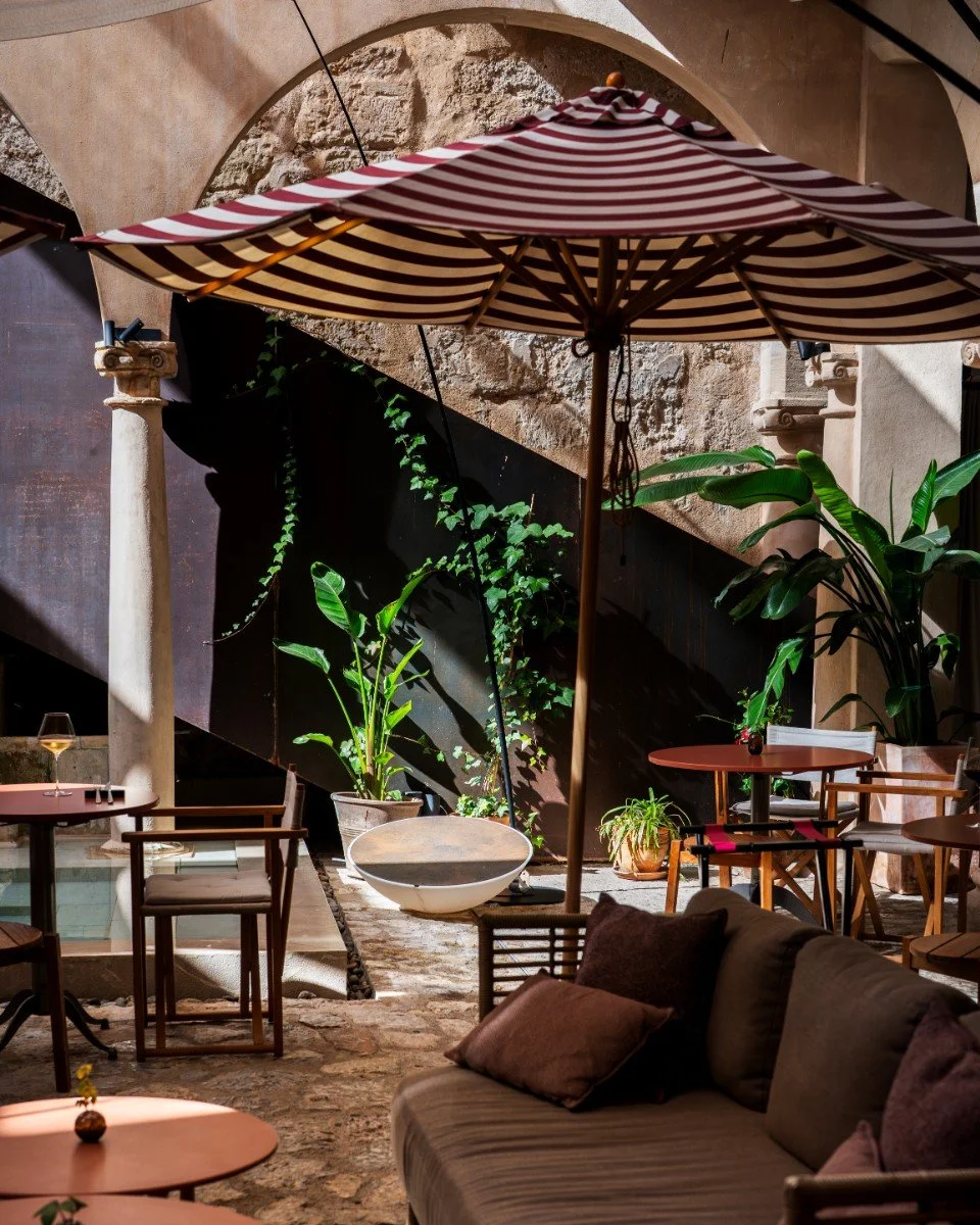 Outdoor patio with striped beach umbrella, potted plants, wooden chairs, and a sofa with pillows, set against stone and stucco walls.