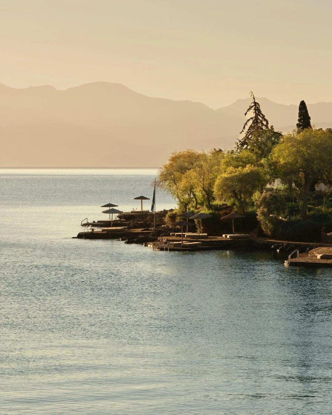 A serene lakeside scene at sunset with trees, lounge chairs, and umbrellas along the shore, mountains in the background.