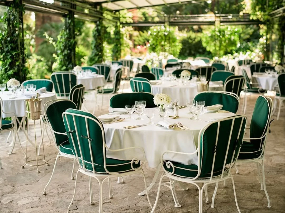 Elegant outdoor dining area with round tables covered in white tablecloths and green upholstered chairs, surrounded by greenery and vines.