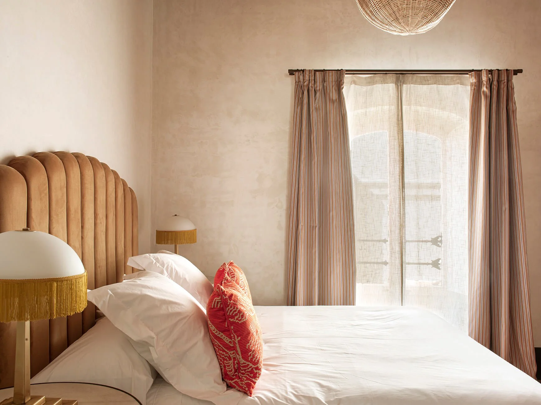A bedroom with a beige headboard, white bed linens, and a red patterned pillow. There are two lamps with fringed shades on either side of the bed. A window with sheer curtains and striped drapes is in the background, letting in natural light.