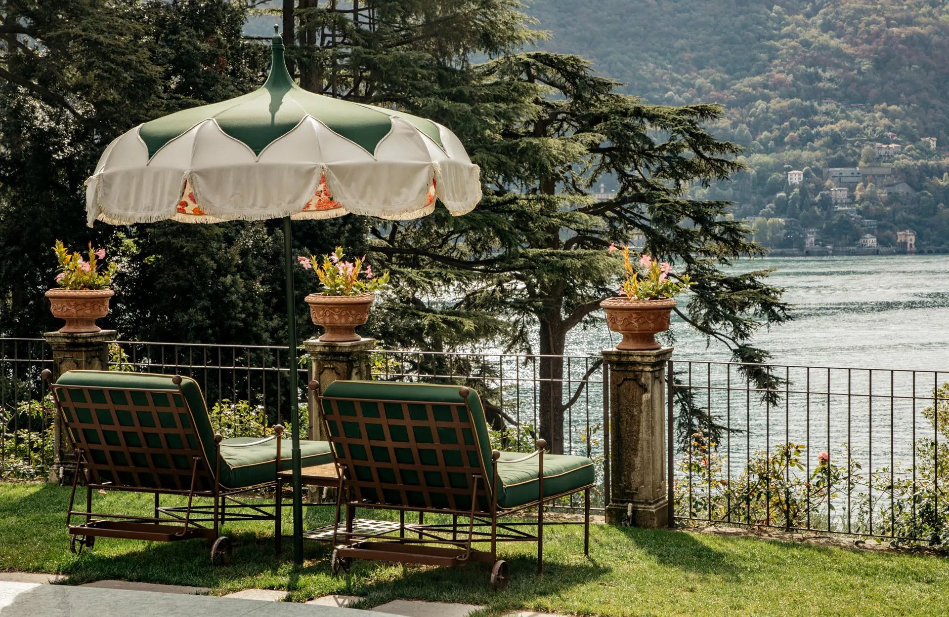 Outdoor patio area with green cushioned chairs, a large white and green umbrella, and potted pink flowers, overlooking a body of water and hillside with buildings in the distance.