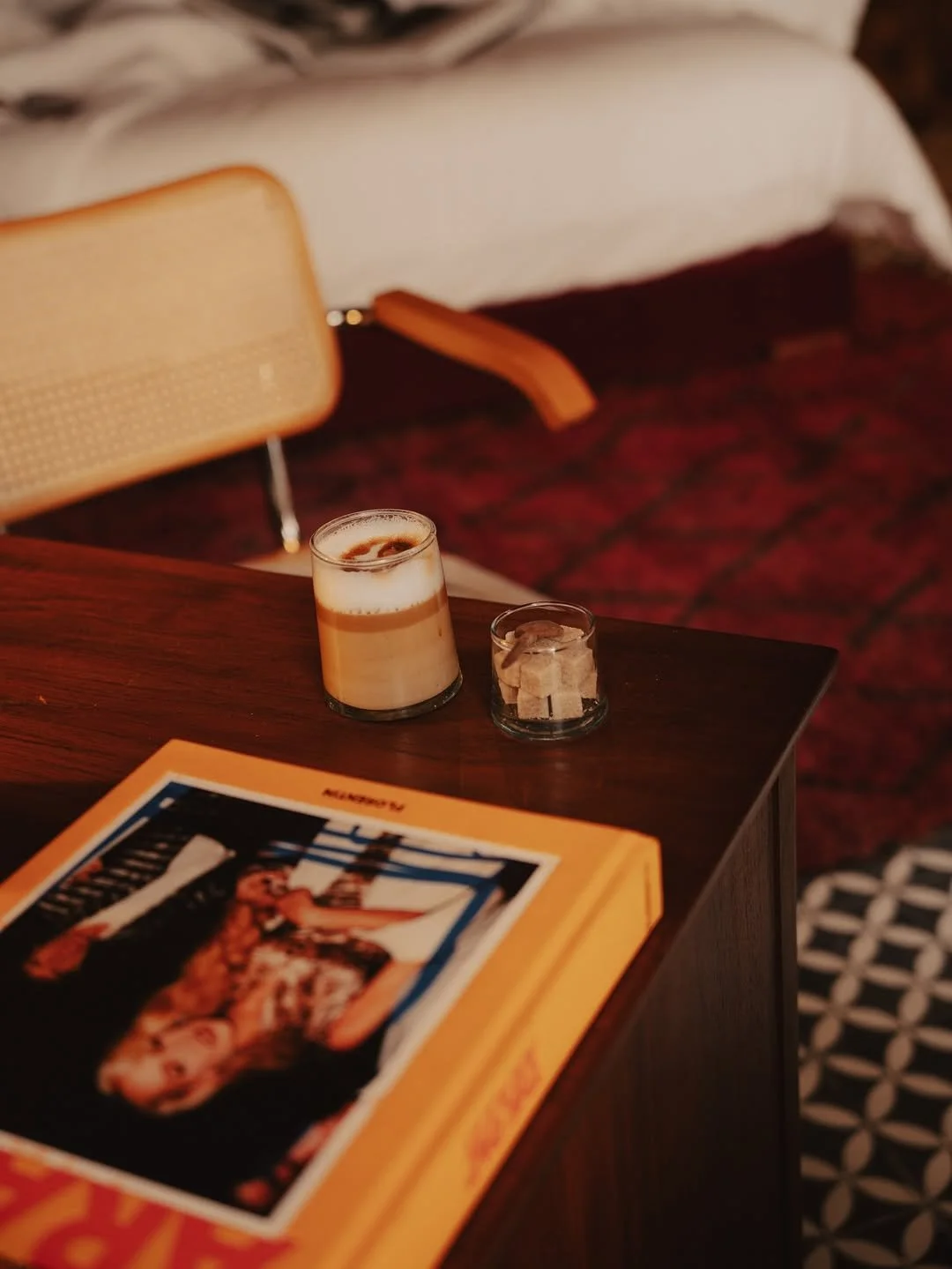 A dark wooden table with a magazine and two drinks, one coffee and one with sugar cubes, in a cozy room with patterned flooring and a beige chair in the background.