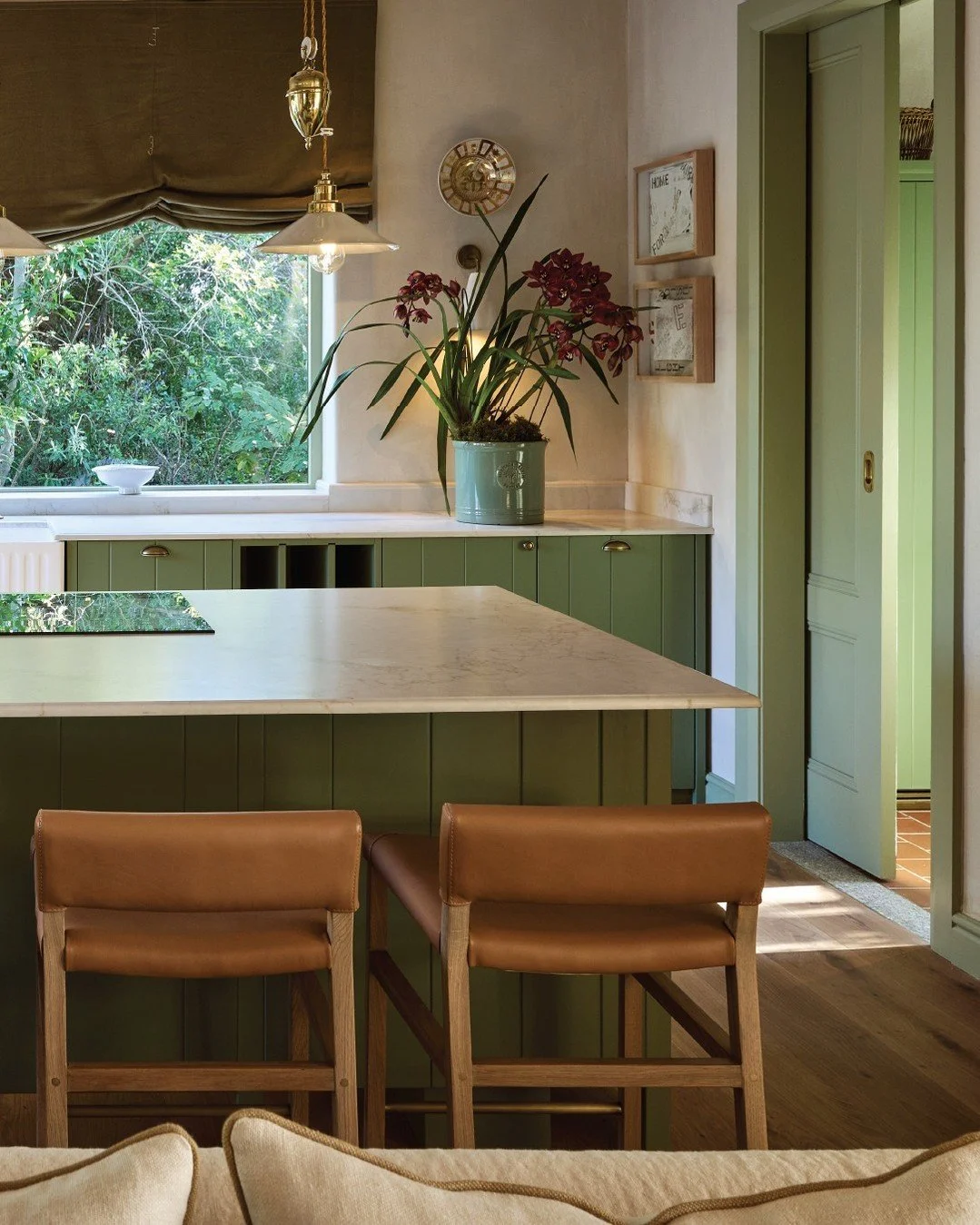 A kitchen with green cabinets, a white marble countertop, a potted orchid on the counter, and a window overlooking greenery outside. Two brown chairs are in the foreground.