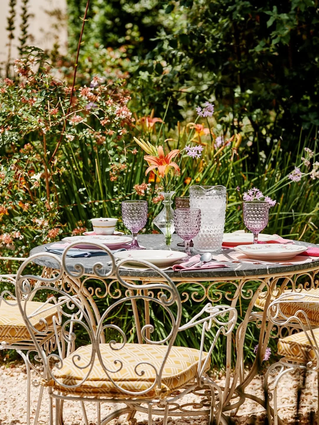Outdoor dining table set with plates, pink glassware, a glass vase with lilies, and a large white textured vase, surrounded by greenery and pink flowers