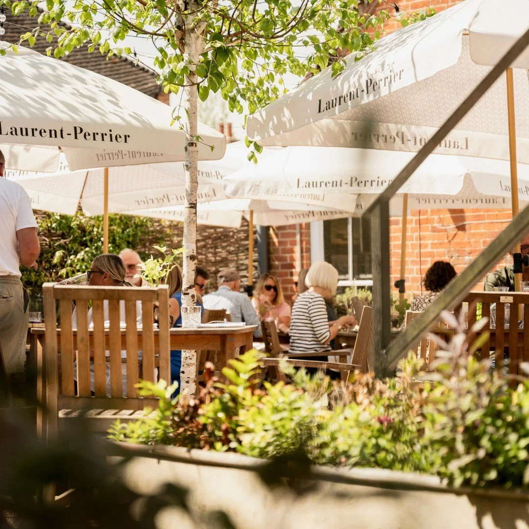 Outdoor patio with wooden tables and chairs, large Laurent-Perrier umbrellas, and people sitting and socializing. There is a tree with green leaves, a brick building in the background, and potted plants in the foreground.