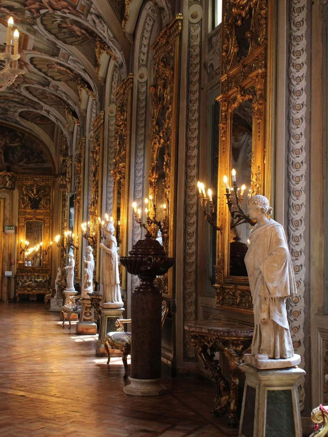 A grand, ornate hallway with gold-gilded mirrors, detailed wall carvings, marble statues, and elegant chairs, illuminated by elaborate wall sconces with candles.