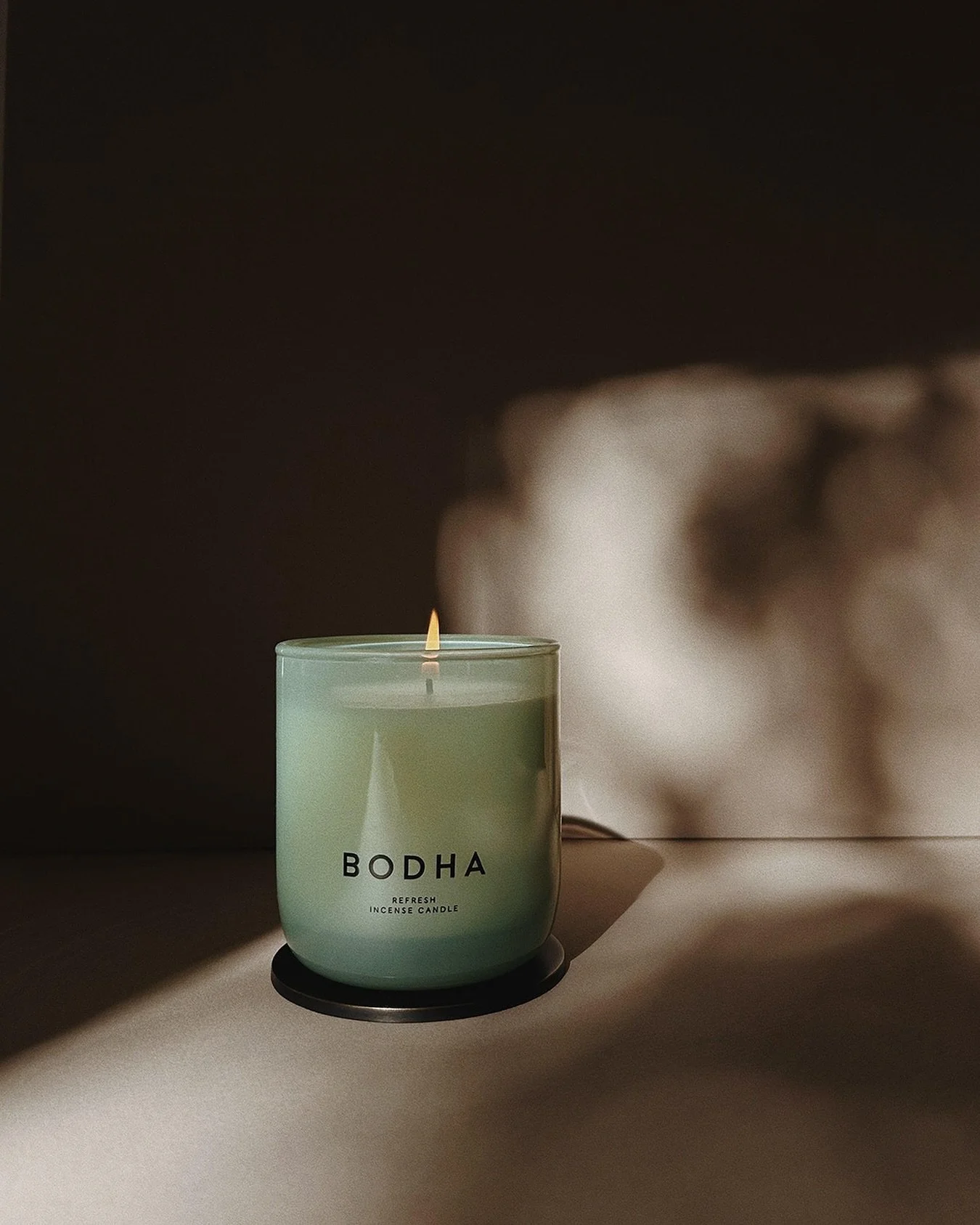 A lit green incense candle with the label 'BODHA Refresh Incense Candle' placed on a surface, casting shadows and a soft glow against a dark backdrop.