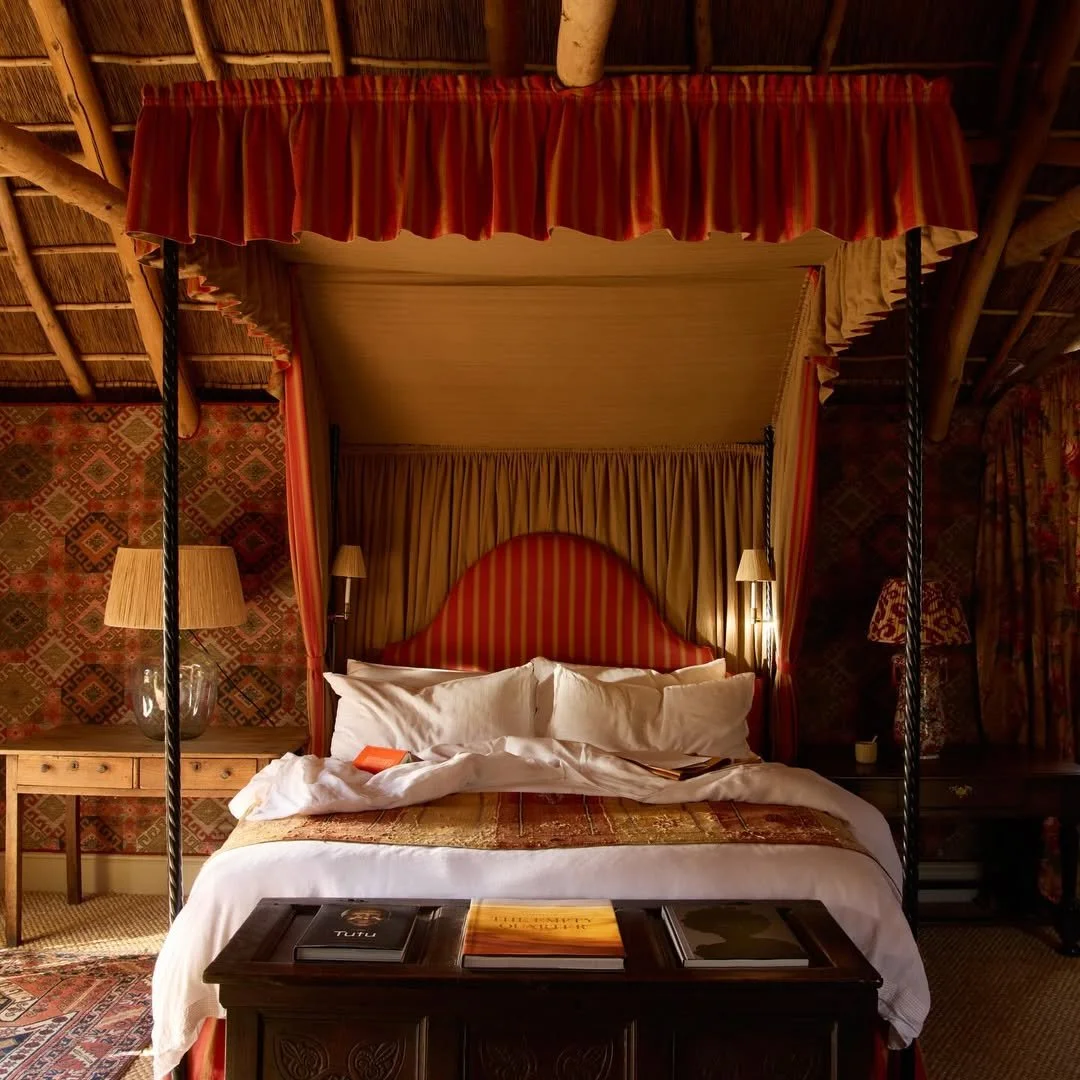A luxurious bedroom with a four-poster bed draped with red and gold curtains, against a patterned wall with lamps on each side, and books on a dark wooden bench at the foot of the bed.