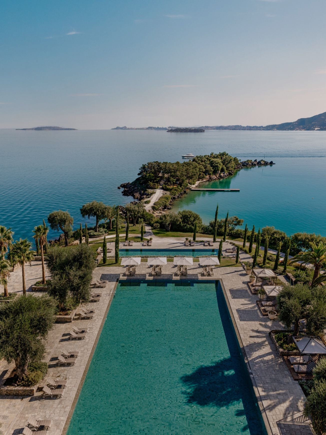 A large rectangular swimming pool surrounded by lounge chairs and umbrellas, with lush trees around it. Beyond the pool, there is a view of a calm body of water with a small island and distant shoreline under a clear sky.