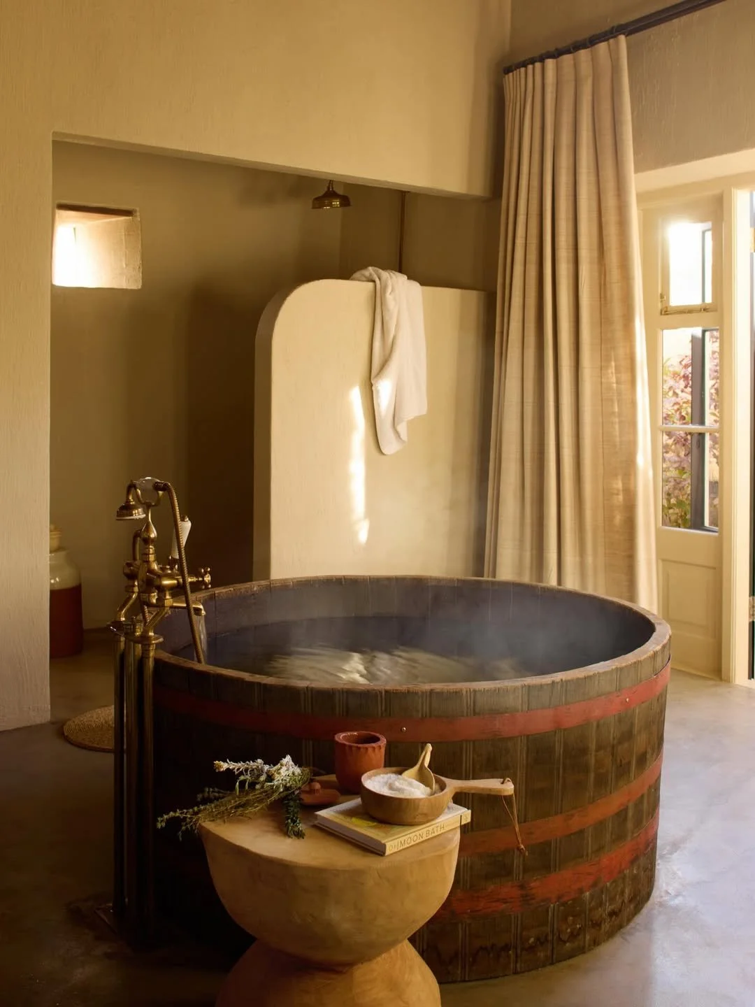 Wooden bathtub filled with hot water in a cozy bathroom with natural light, beige curtains, and spa items on a wooden stool.