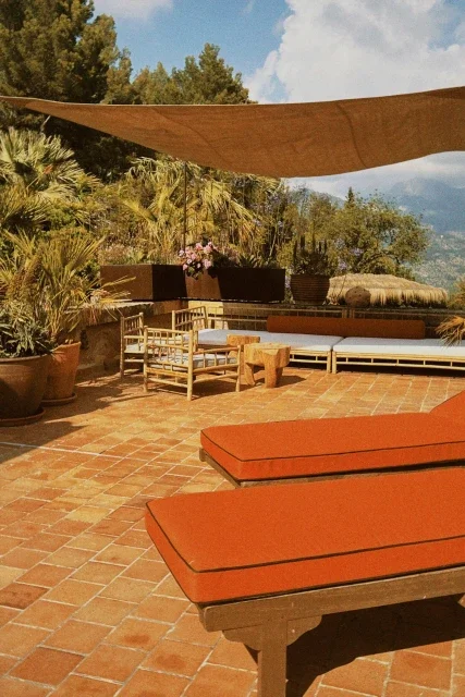 Outdoor patio with orange lounge chairs, wooden bench, potted plants, and a shaded canopy, overlooking a scenic landscape with trees and mountains.