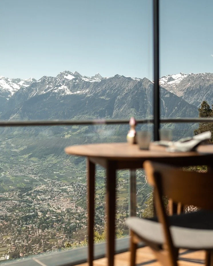 A scenic view of snow-capped mountains and a valley seen from inside a room with a wooden table and chairs, large window with a black frame.