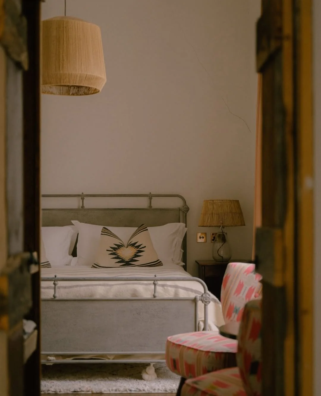A cozy bedroom seen through a doorway, featuring a gray metal bed with white linens and a decorative pillow with a geometric pattern. There are two yellow table lamps, one on a bedside table and another hanging from the ceiling, and a pink patterned chair in the foreground.