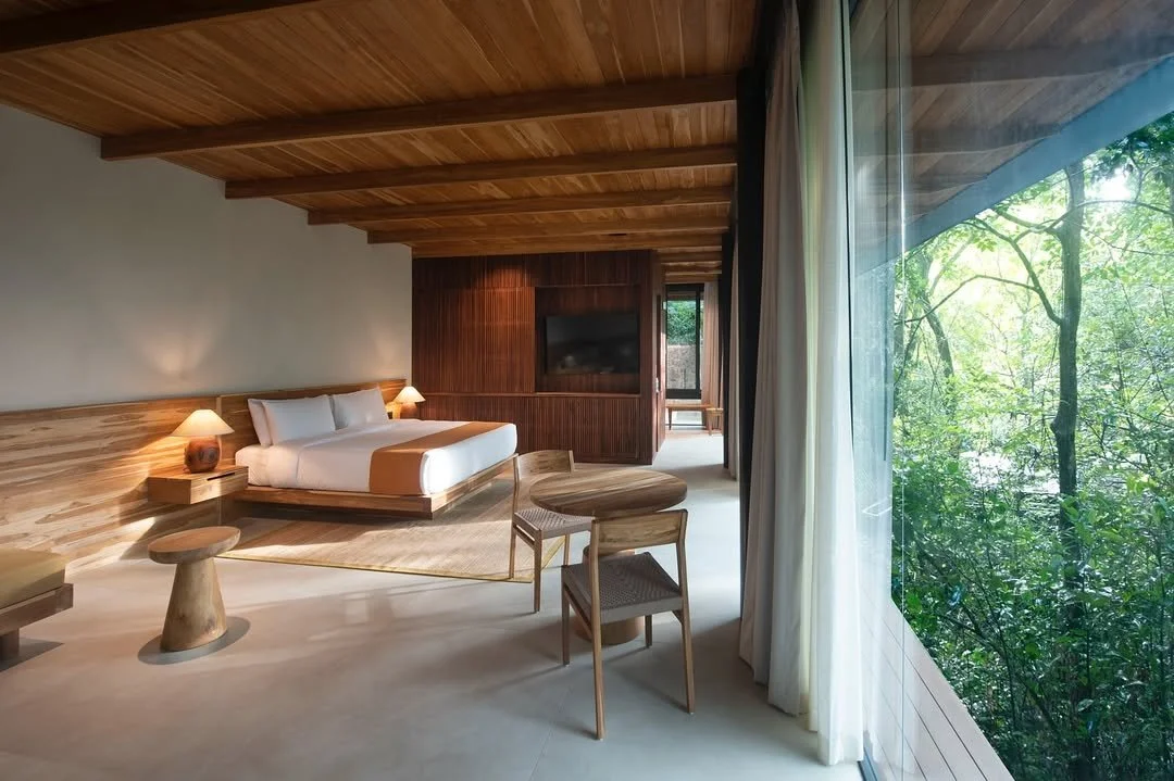 A modern bedroom with a wooden ceiling, a bed with white bedding and bedside lamps, a small round table with a chair, and large glass windows showing green trees outside.