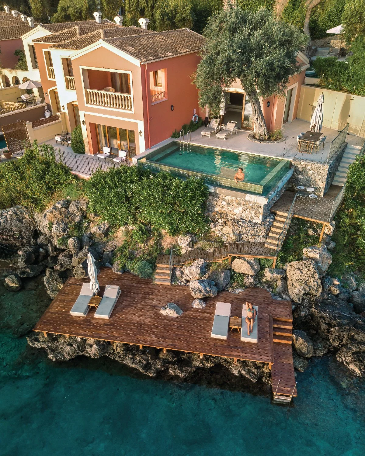 Aerial view of a luxurious seaside house with a swimming pool, surrounded by trees and rocks. The house features multiple levels with outdoor seating, a private pool with a person relaxing in it, and a large wooden deck extending over the water with sun loungers and umbrellas.