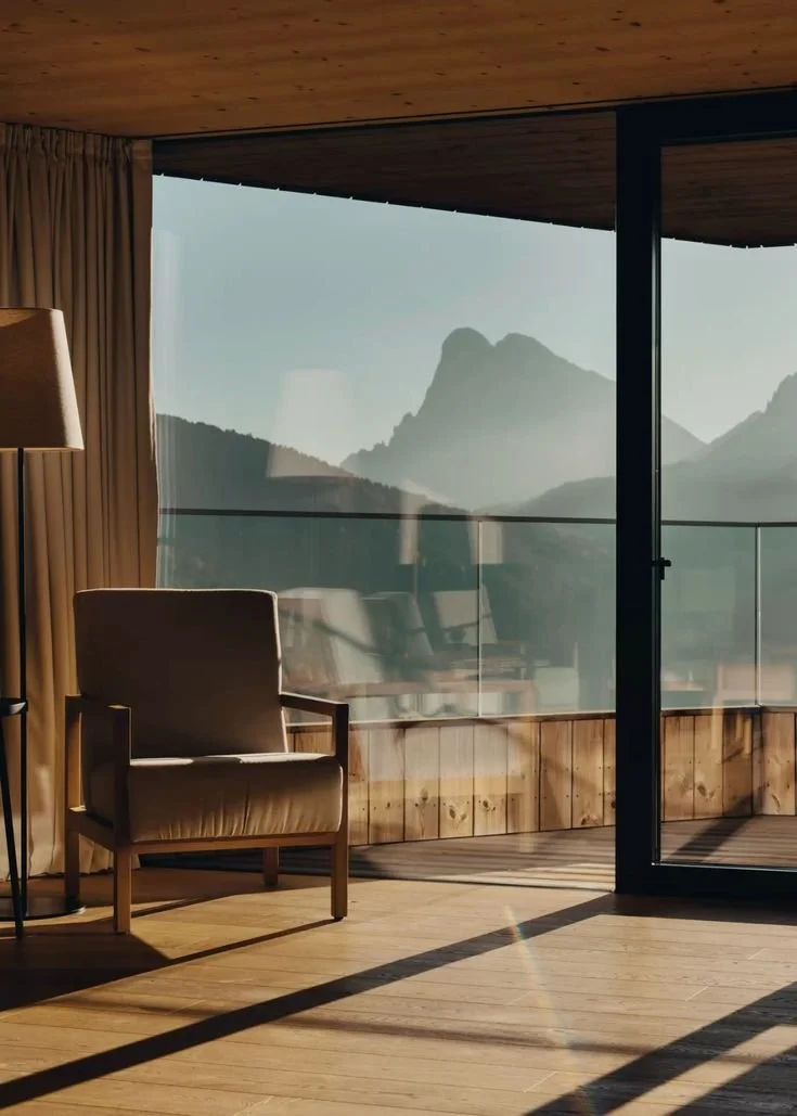 Room with a wooden floor, beige armchair, standing lamp, and glass balcony doors overlooking mountains.