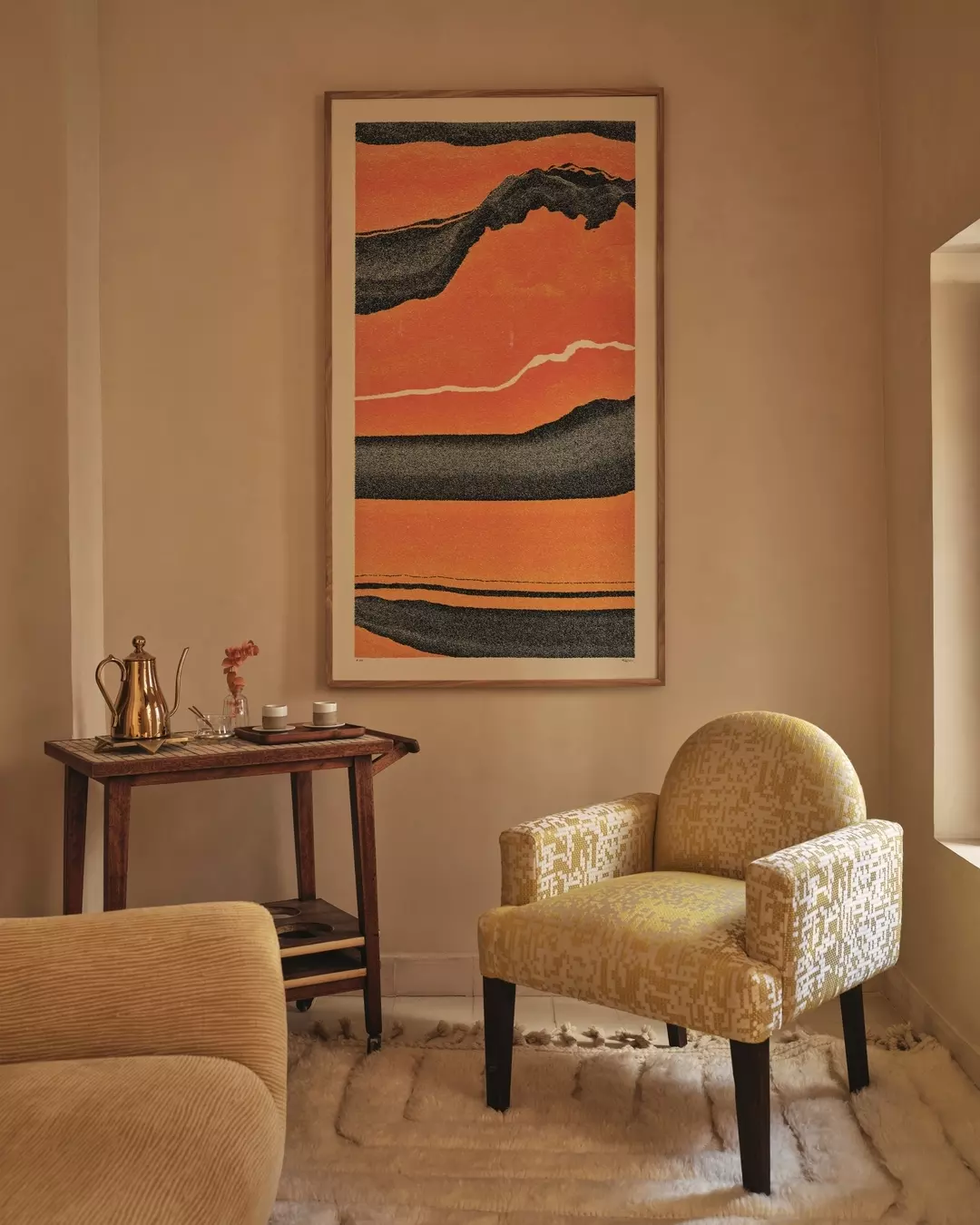 A cozy living room corner with a beige armchair featuring a geometric pattern, a wooden side table with a gold teapot, two cups, and pink flower, and a large abstract picture with black, orange, and white colors on the wall.