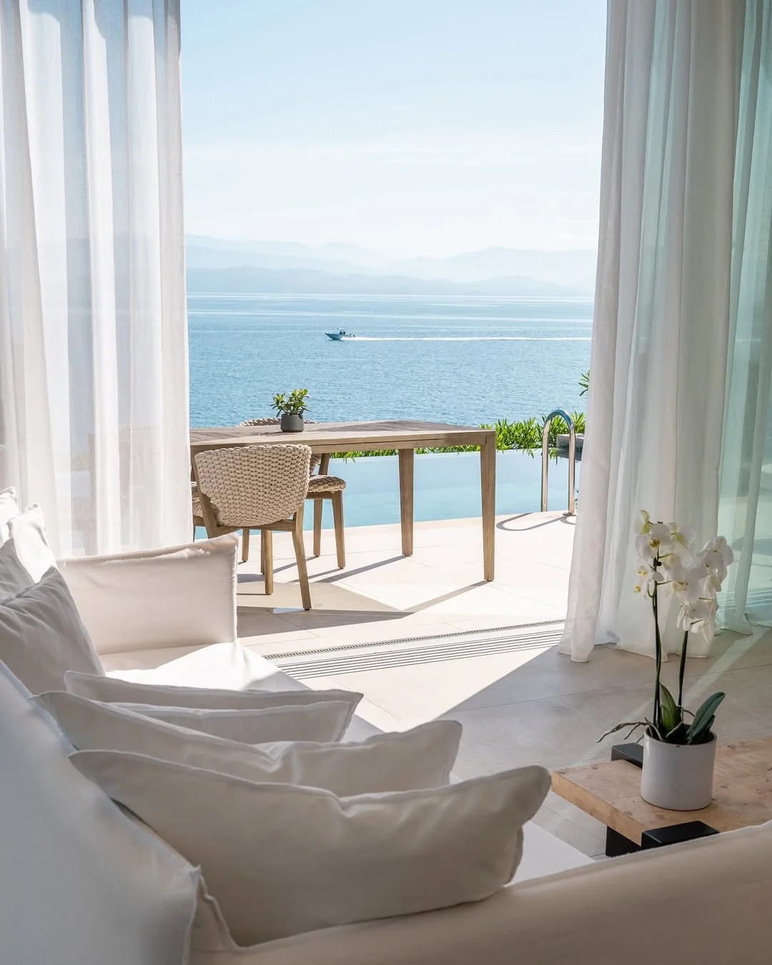 A bright, airy room with white curtains, a white sofa with pillows, a wooden side table with a white orchid, and a sliding glass door leading to a balcony overlooking the ocean with a boat in the distance.