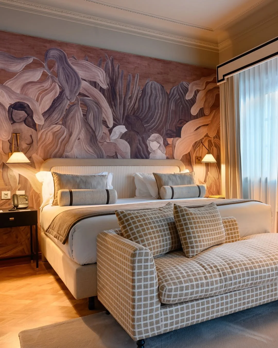 Elegant hotel bedroom with a large bed, decorative pillows, a patterned sofa, wall mural with abstract floral design, bedside lamps, and window with curtains.