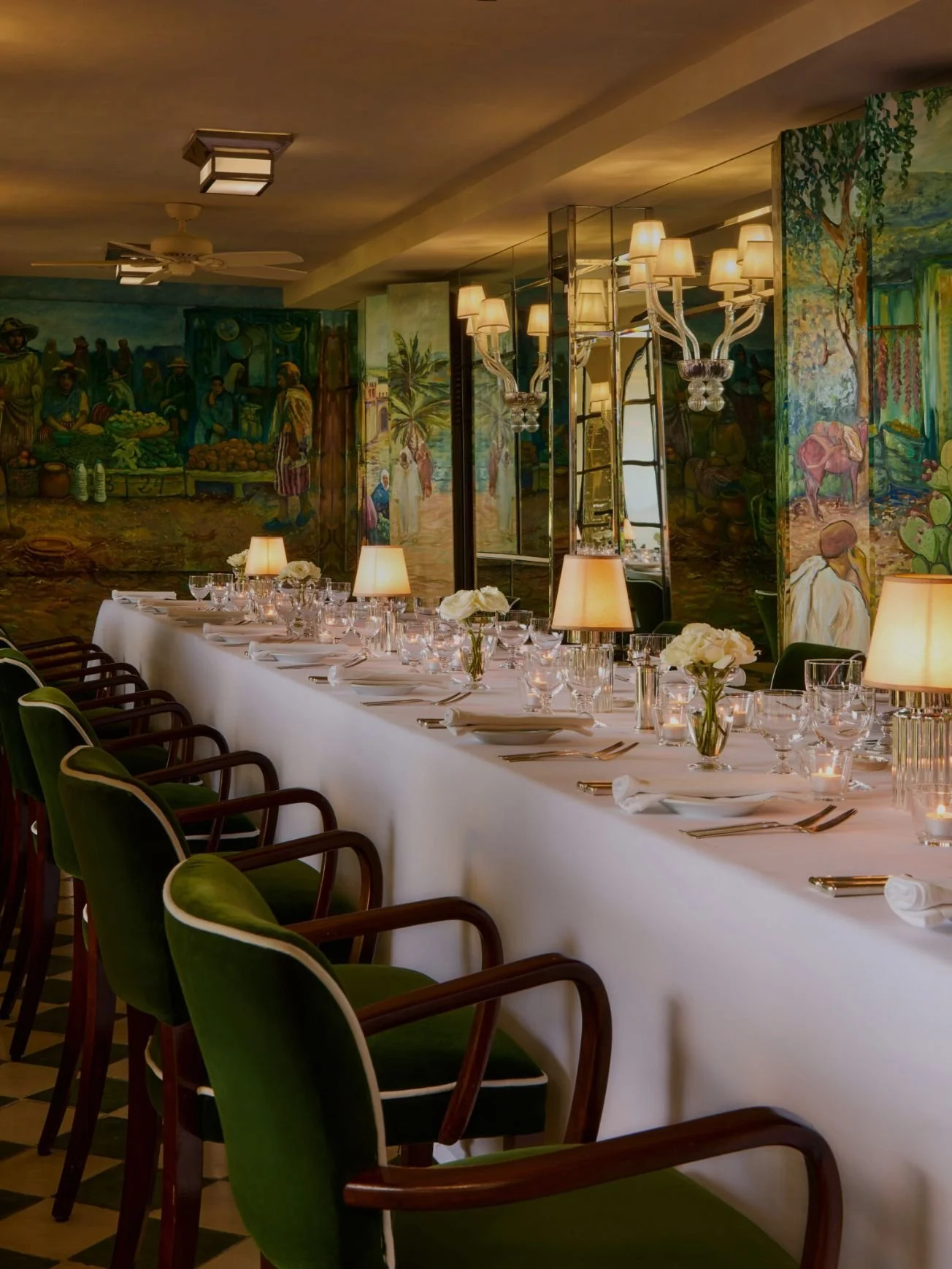 A long dining table set with white tablecloth, wine glasses, and small flower arrangements, surrounded by green velvet chairs with wooden arms. The room has ornate wall lamps, a mirrored wall, and vibrant murals depicting people, nature, and rural scenes.
