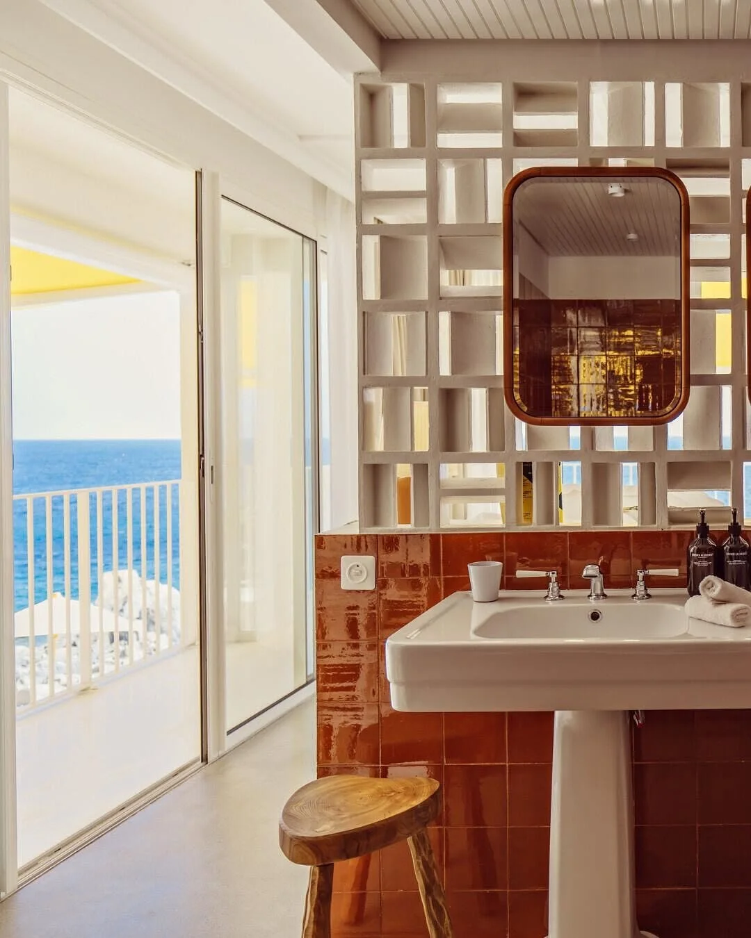 Bathroom with a view of the ocean through a sliding door, featuring a pedestal sink, a small wooden stool, and a decorative wall partition with a mirror.