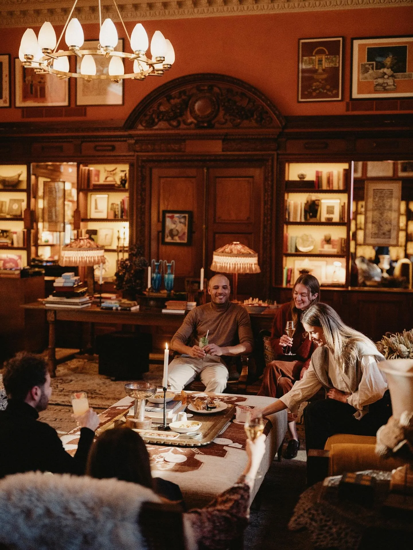 A group of people are gathered in a warmly lit, elegant living room with vintage decor. They are sitting and standing around a table with food and drinks, smiling and engaging in conversation. The background features built-in shelves filled with books and decorative items, and a chandelier hangs from the ceiling.