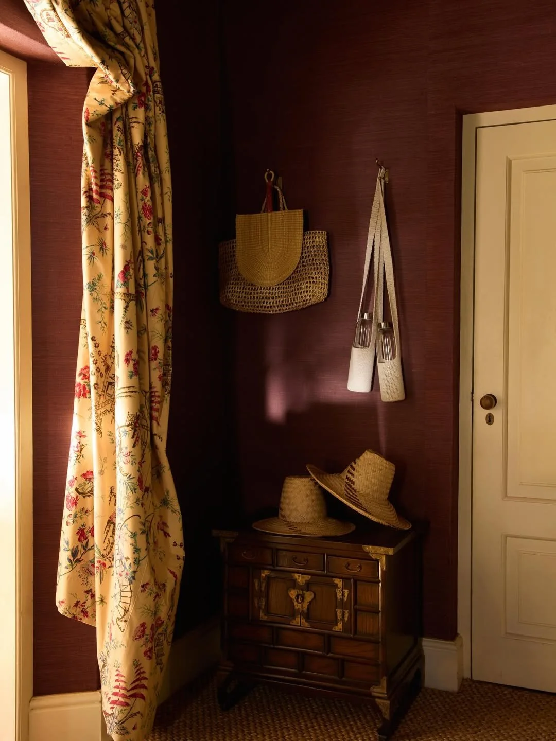 corner of a room with burgundy wallpaper, a closed white door on the right, a floral curtain on the left, a small wooden cabinet with three hats placed on top, a wicker basket, and two hanging fabric pouches or bags on the wall.