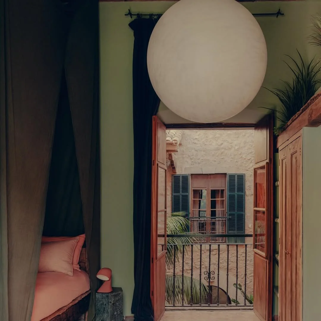 View from a bedroom looking out a balcony with open wooden shutters, stone building with windows and green shutters, potted plants on the balcony.
