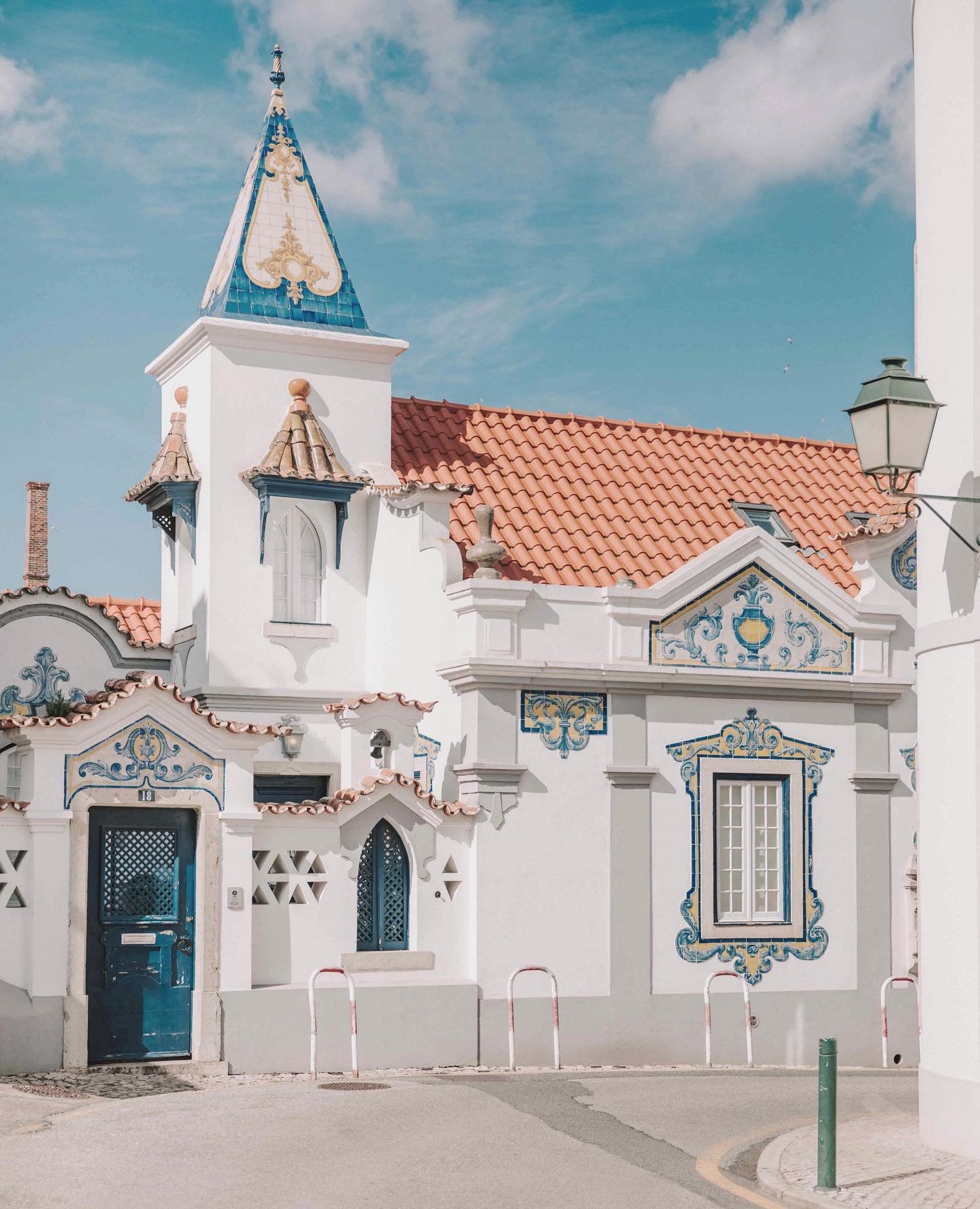 A white building with ornate blue and gold decorative elements, red-tiled roof, and a steeple with blue tiles and gold embellishments, under a partly cloudy sky.