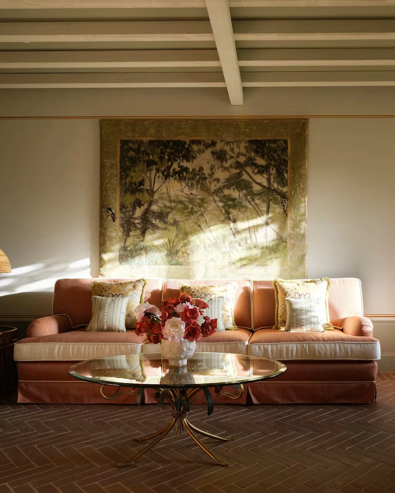 A living room with a pink sofa decorated with patterned cushions, a glass coffee table with a vase of pink and white flowers, a large landscape painting with trees and cranes behind the sofa, and sunlight casting shadows on the wall.