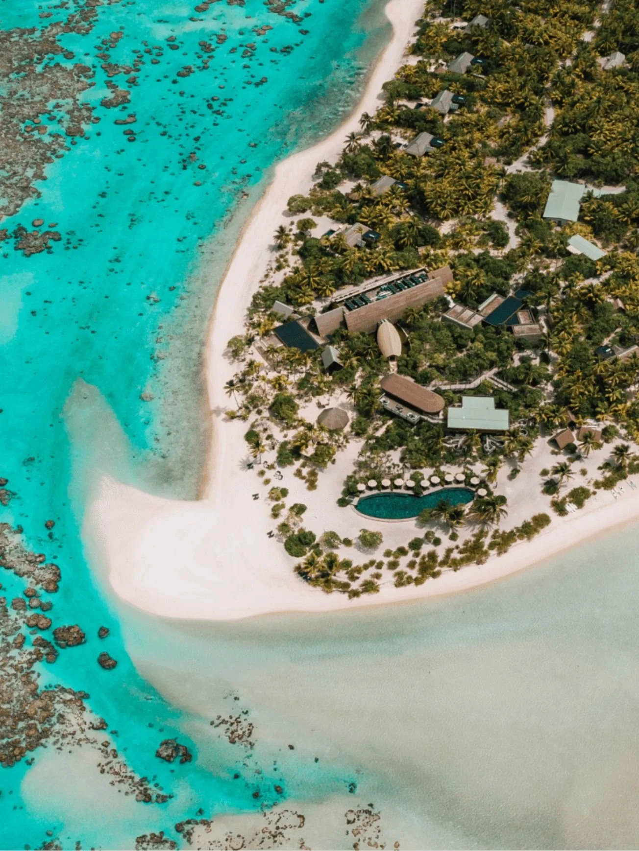 Aerial view of a luxurious tropical island resort with white sandy beaches, turquoise waters, lush palm trees, and multiple buildings including a pool with sun loungers.