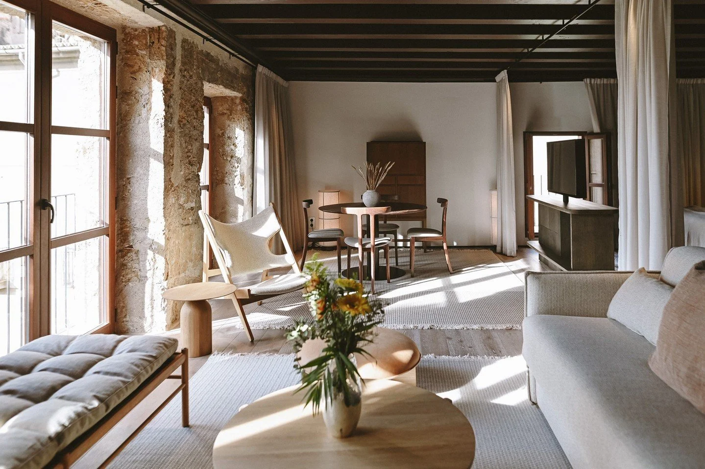 Living room with large windows, exposed brick wall, beige sofa, wooden furniture, and a round dining table with chairs, decorated with a vase of flowers.