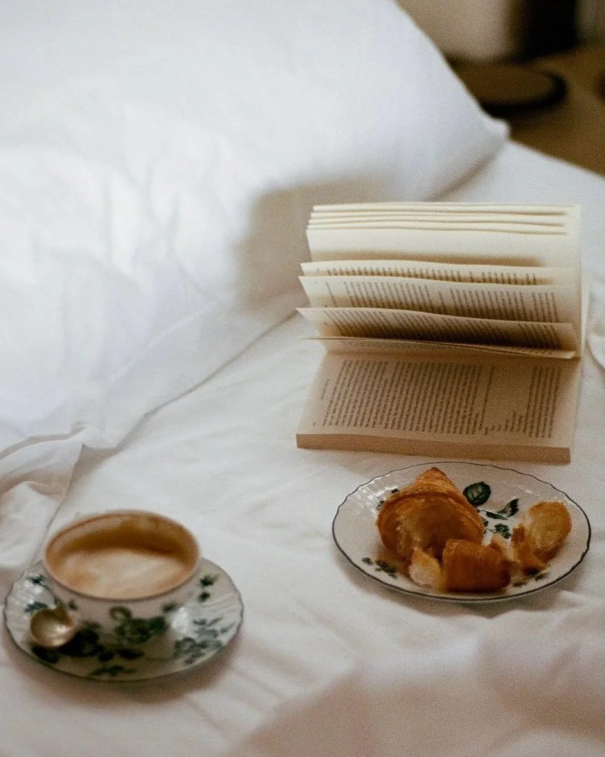 A cup of coffee with a small spoon on a decorative saucer, a plate with a croissant and some food pieces, and an open book on a white bed with a pillow.