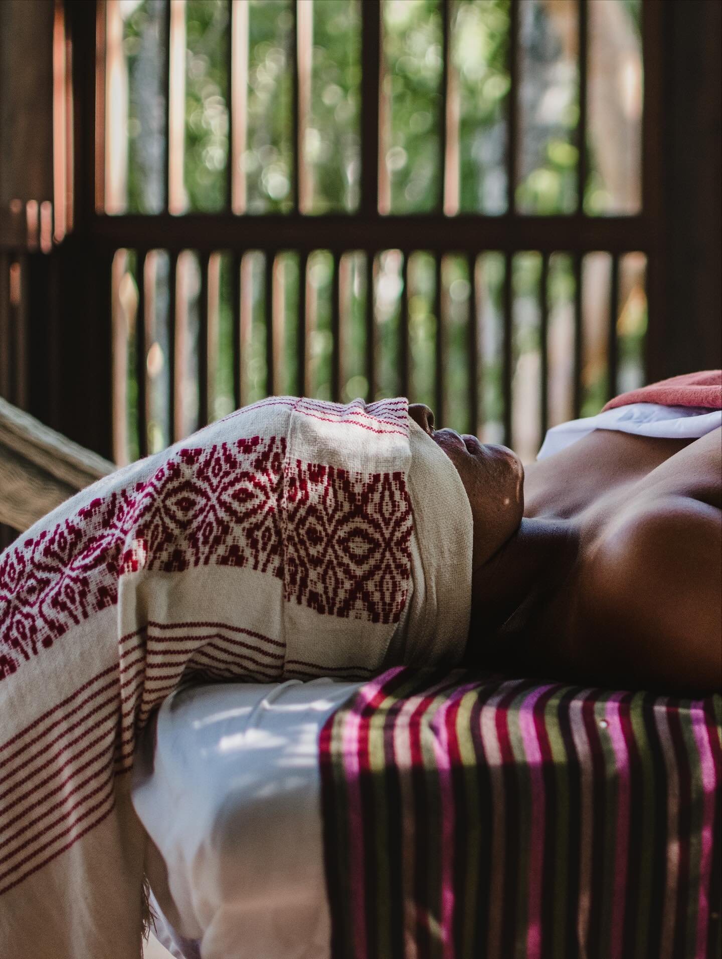 A person lying down inside a structure with wooden bars, covered with a towel or blanket with a red geometric pattern, and wearing striped clothing, with greenery visible outside through the wooden slats.