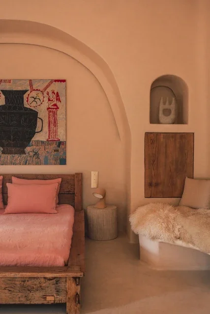 A bedroom corner with a bed featuring pink bedding and pillows, a side table with a small lamp, and a cozy seating area with a fluffy white throw and a beige pillow. Artworks and decorative niches adorn the wall.