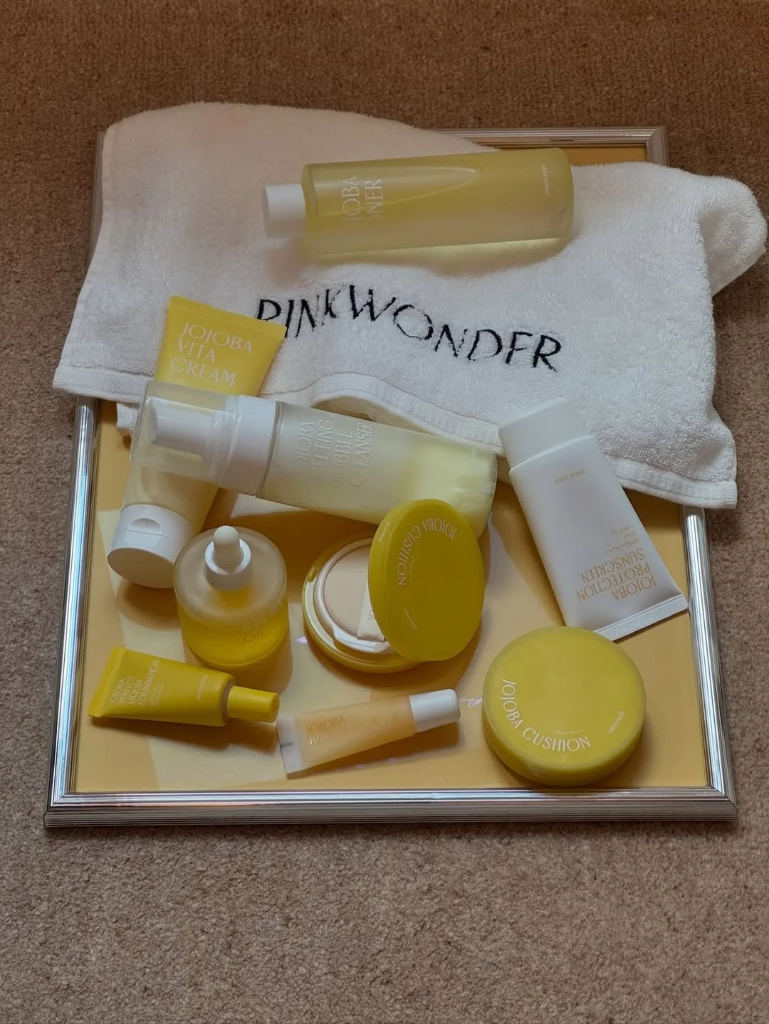 Collection of skincare and beauty products with yellow accents on a tray, including creams, serums, and lotions, with a white towel and a mirror at the back.