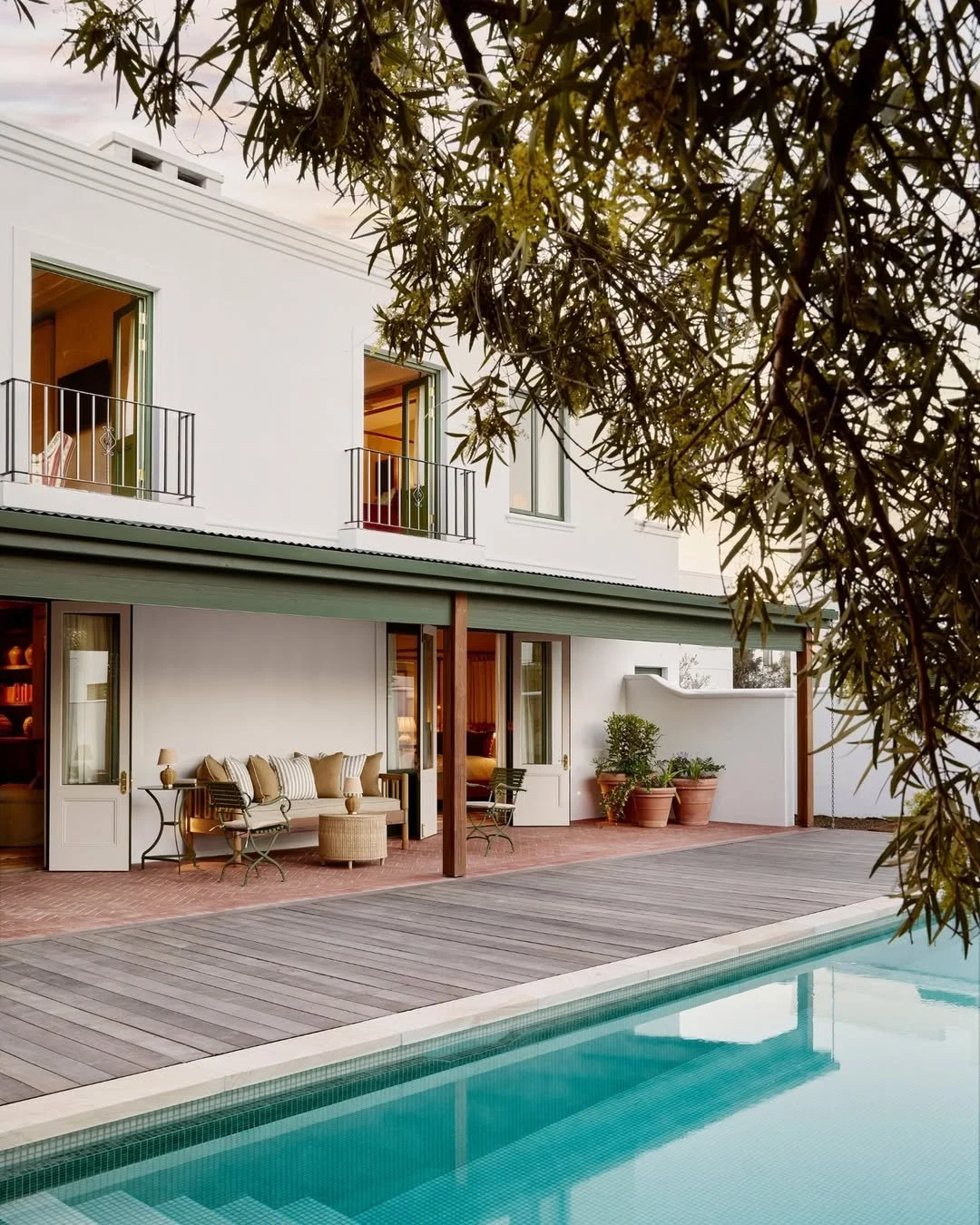 A modern white house with balconies, outdoor seating area with cushions, and a swimming pool in the foreground, framed by tree foliage.