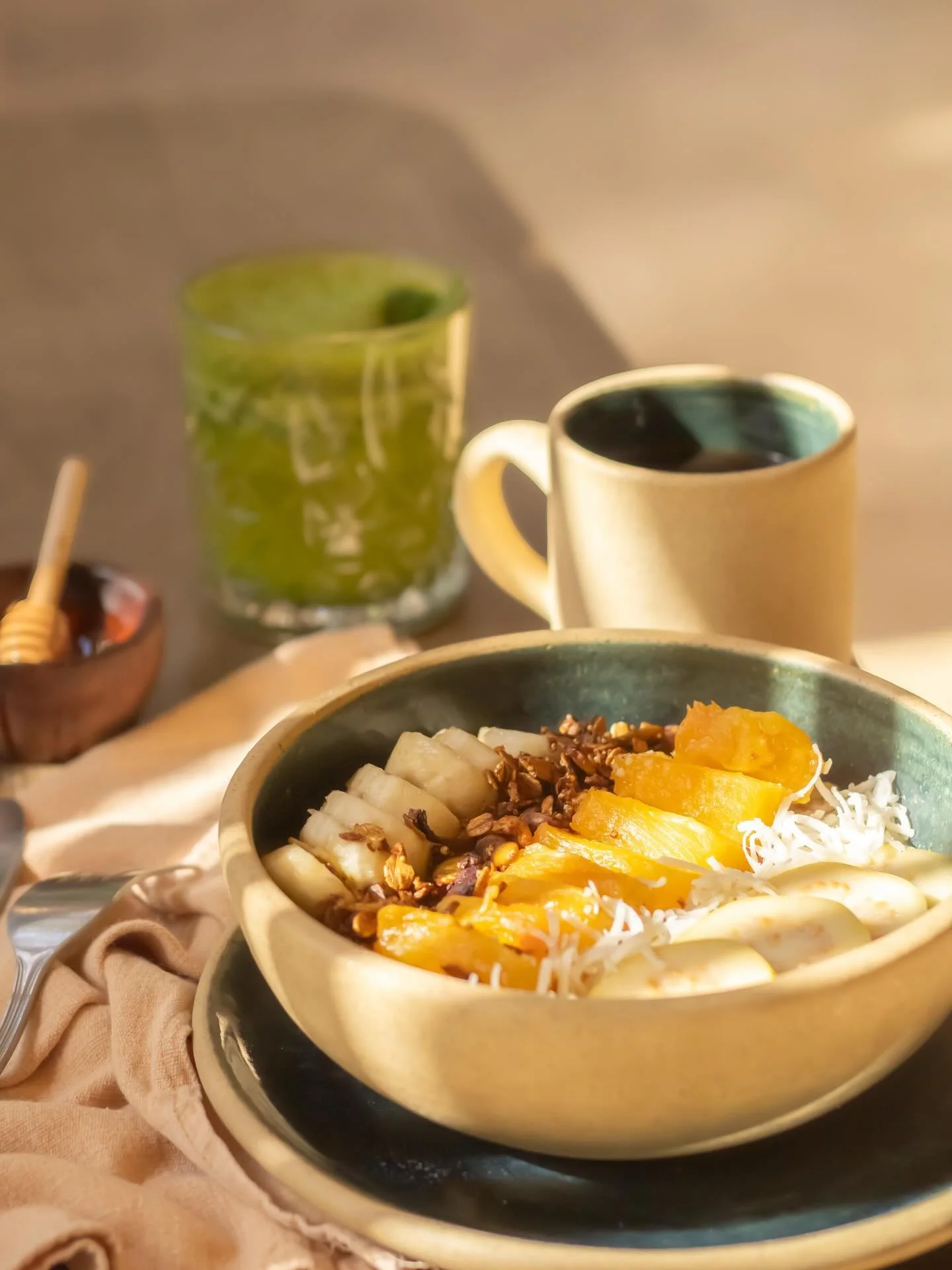 A bowl of yogurt topped with sliced bananas, pineapple chunks, shredded coconut, and granola. In the background, a green container of honey, a beige cup of coffee, and a small jar of honey with a honey dipper are visible on a beige cloth.
