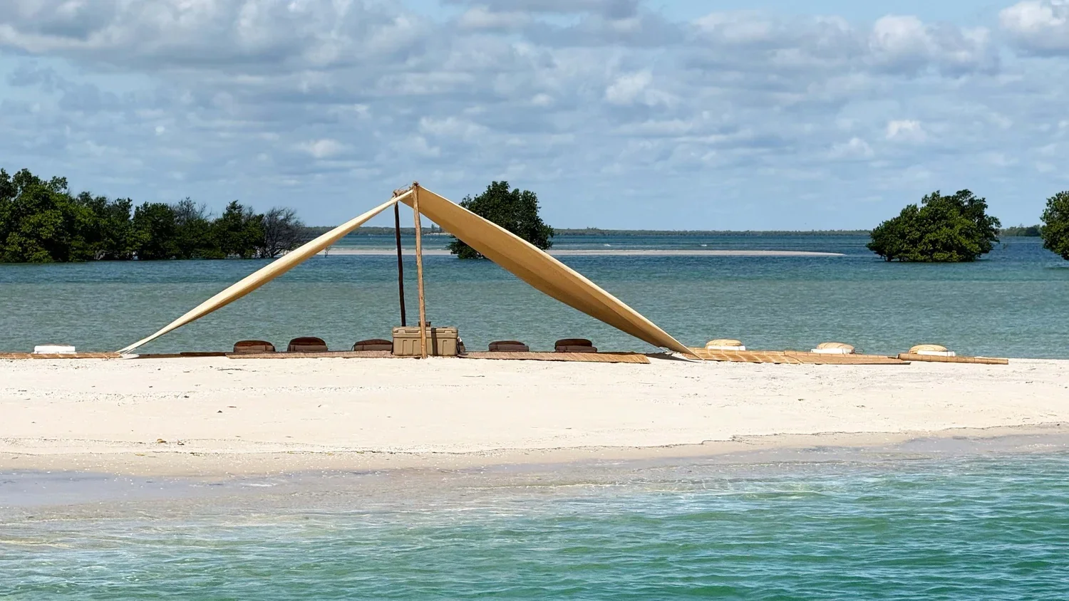 A beach scene with a sailboat sail set up on the sandy shore, amid calm turquoise waters and a backdrop of green trees against a partly cloudy sky.