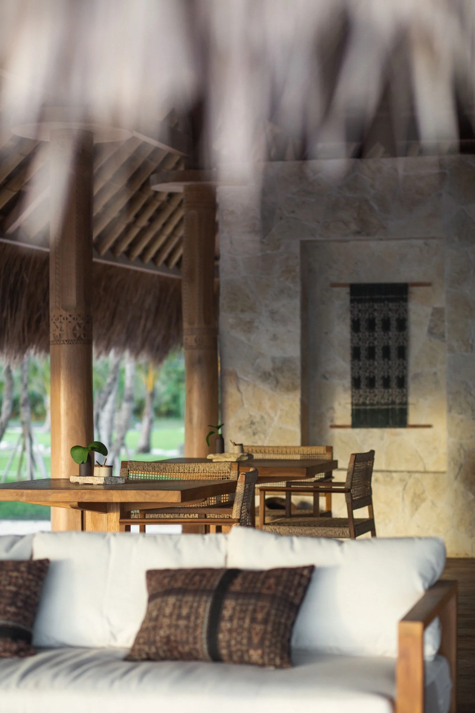 A cozy outdoor seating area with a round wooden table set with a small potted plant, surrounded by wooden chairs with woven seats, under a thatched roof with wooden beams, overlooking a grassy yard with trees.