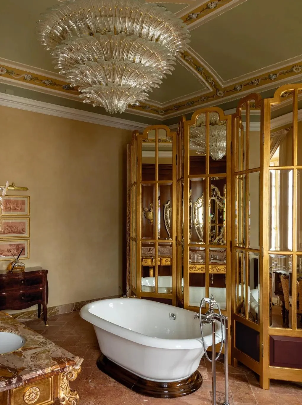 Luxurious bathroom with a freestanding white bathtub, ornate wooden cabinetry, a large glass chandelier, and a decorative folding mirror screen.