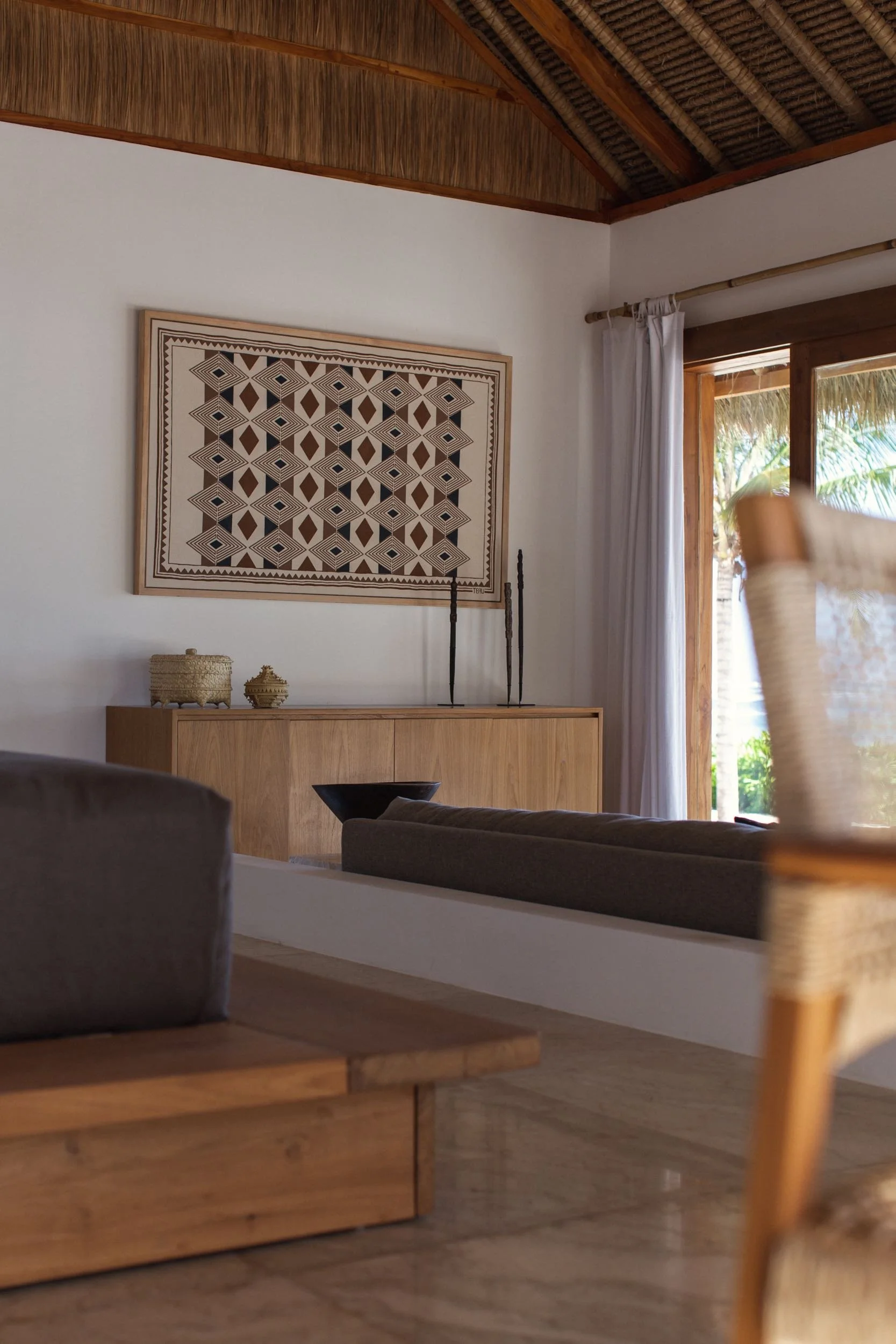 Interior view of a room with a wooden sideboard, decorative objects, a patterned wall hanging, curtains, a sliding glass door, and tropical outdoor scenery.