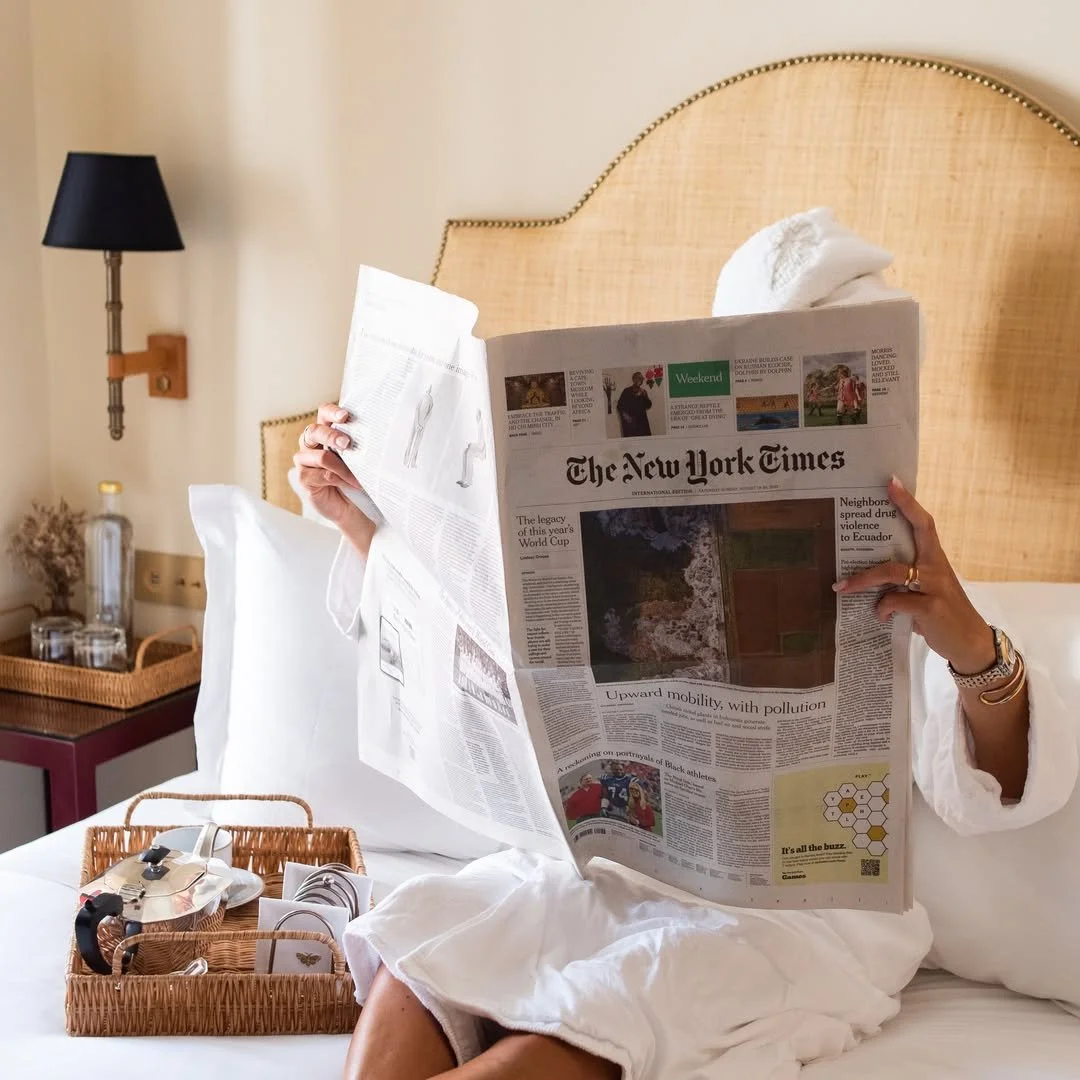 Person reading a newspaper in bed, with a basket of toiletries and a bedside lamp nearby.