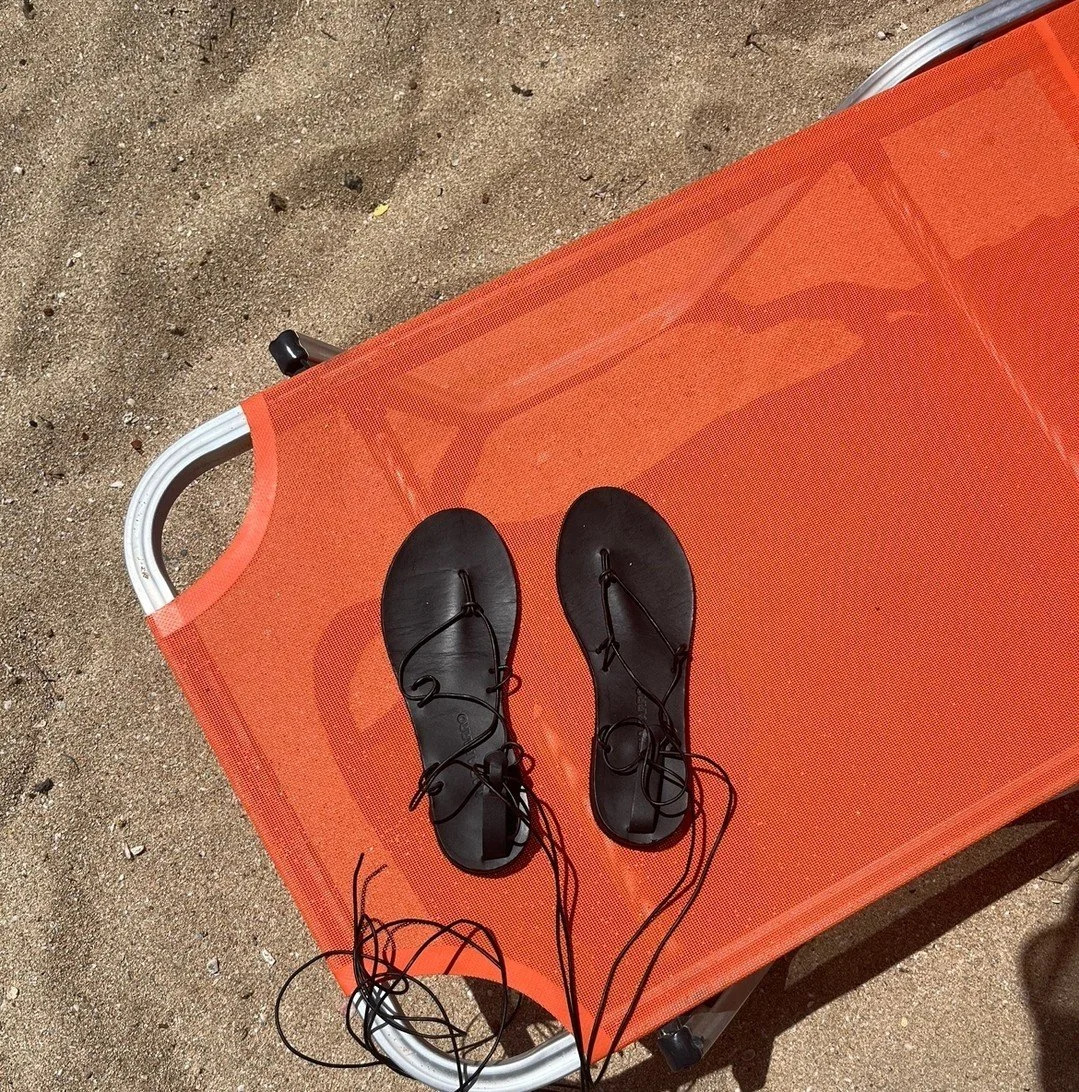A pair of black sandals with ankle straps placed on an orange lounge chair outdoors on sandy ground.