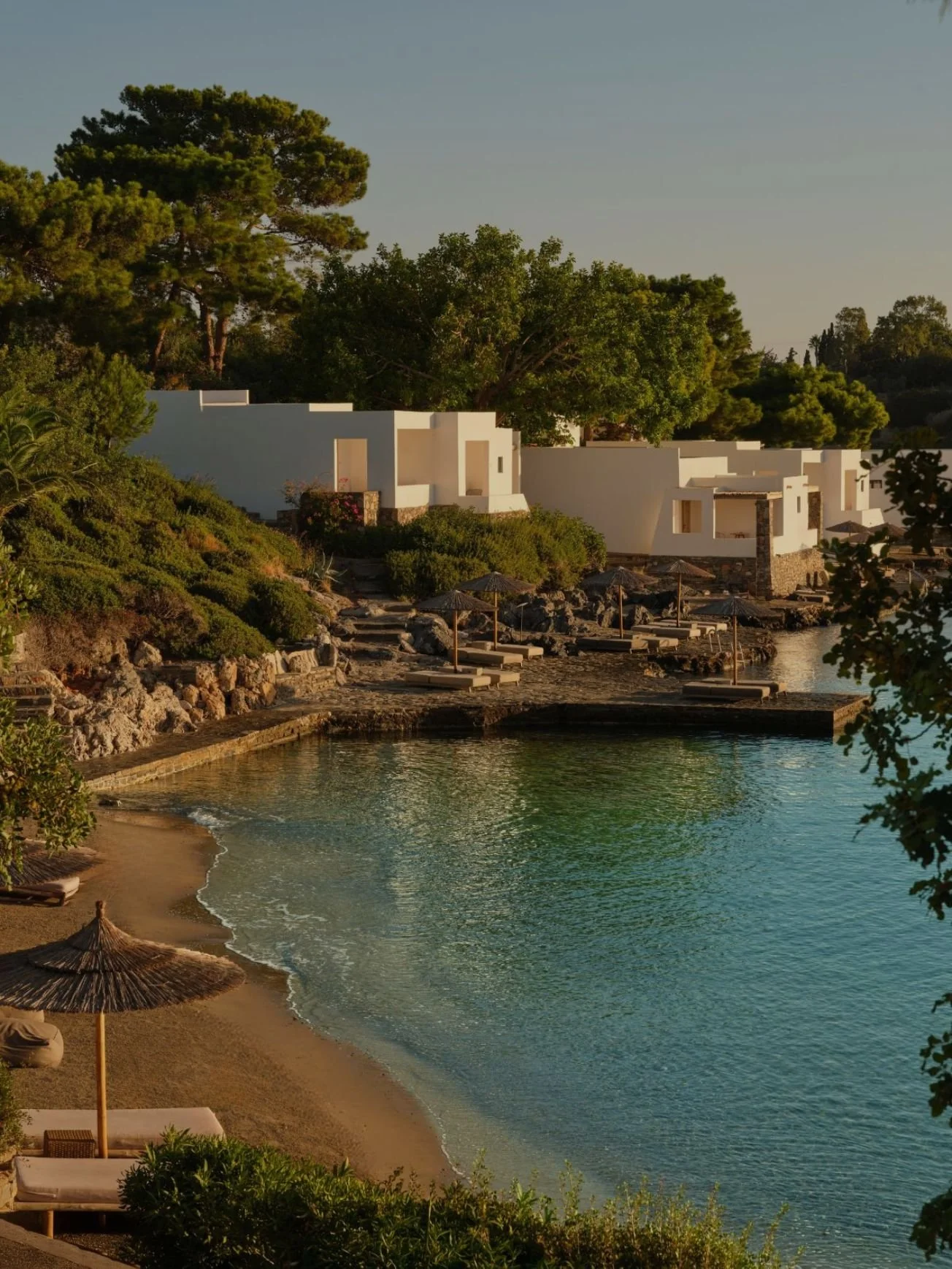 Beach with calm water, lounge chairs with umbrellas, lush green trees, and modern white buildings in the background under a clear sky.