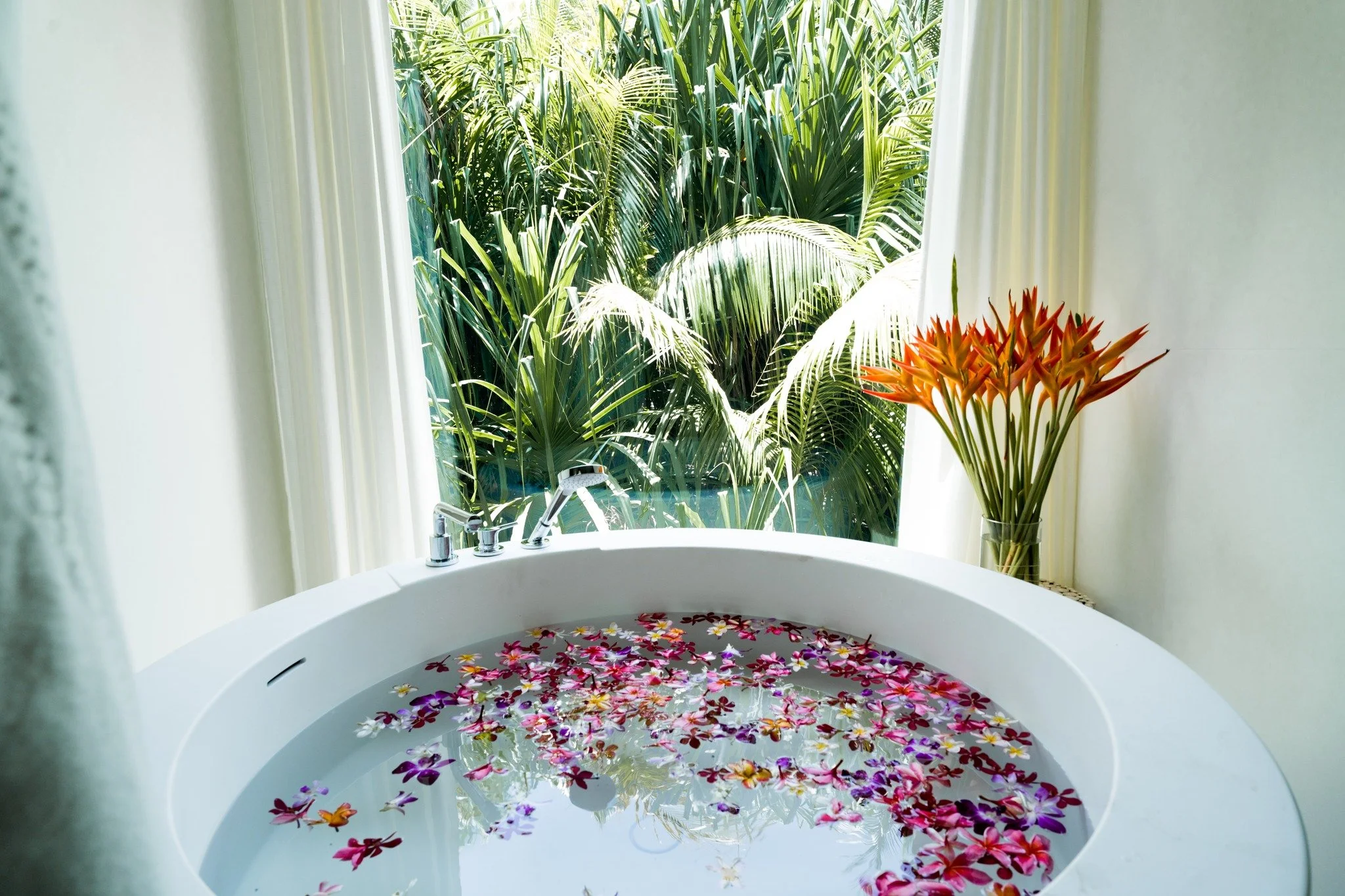 A white bathtub filled with water, decorated with colorful flower petals, situated by a window with lush green tropical plants outside.