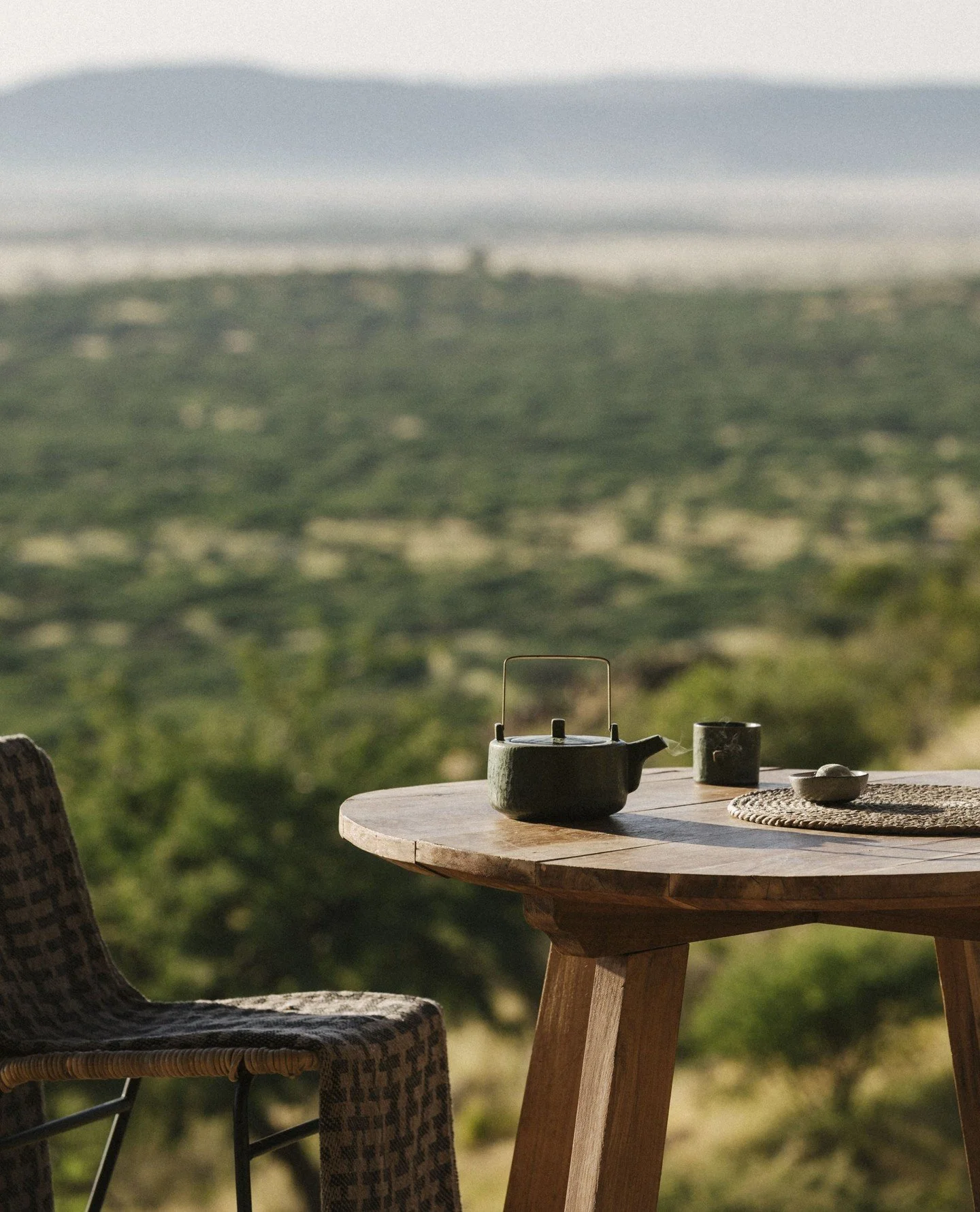 A wooden table with a teapot, a teacup, and a woven placemat, overlooking a lush, green landscape with rolling hills and a cloudy sky.