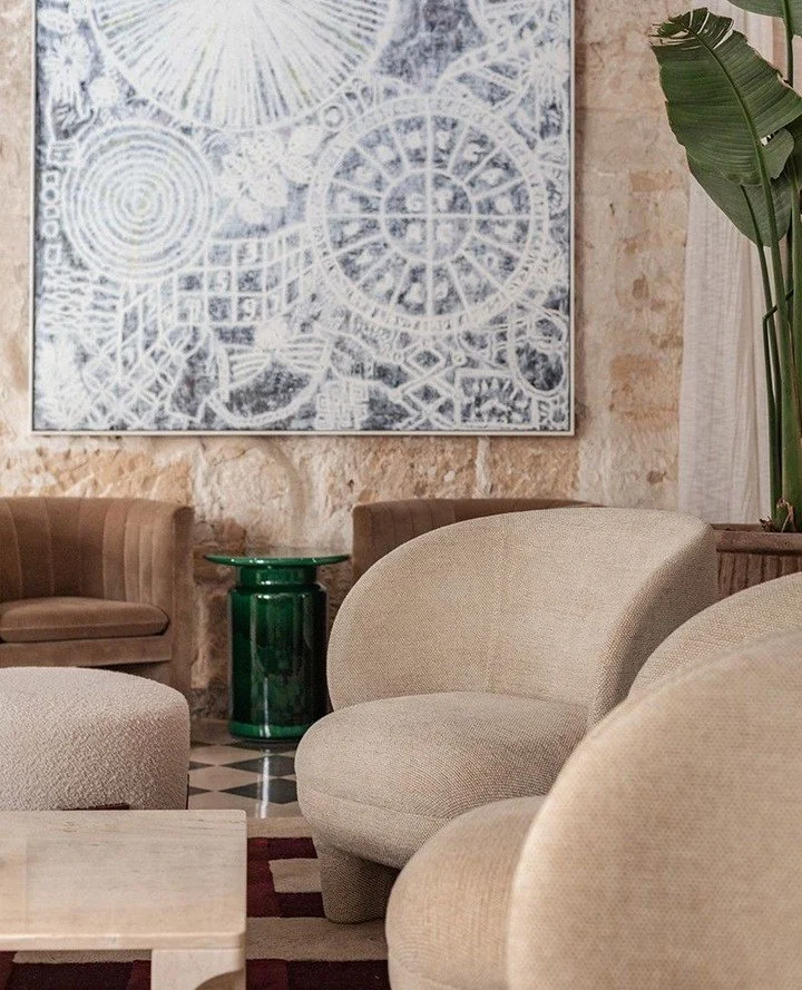 A cozy living room with beige armchairs, a brown sofa, a green cylindrical side table, large green indoor plant, and abstract art on a stone wall.