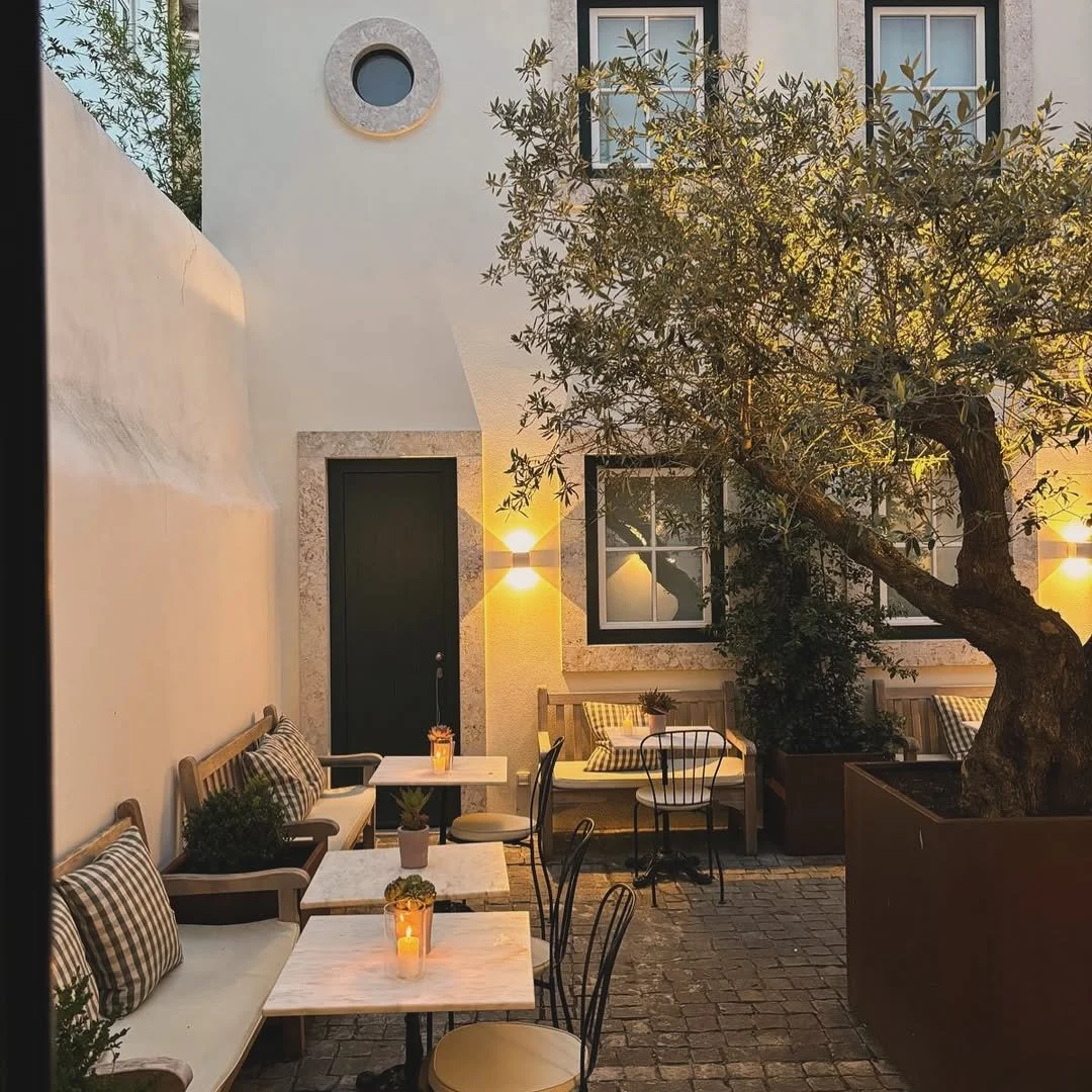 An outdoor patio with seating, including benches with checkered pillows and small tables with candles, next to a large tree with a textured trunk. The patio is adjacent to a white building with a black door, a window, and warm outdoor wall lights, creating a cozy evening atmosphere.