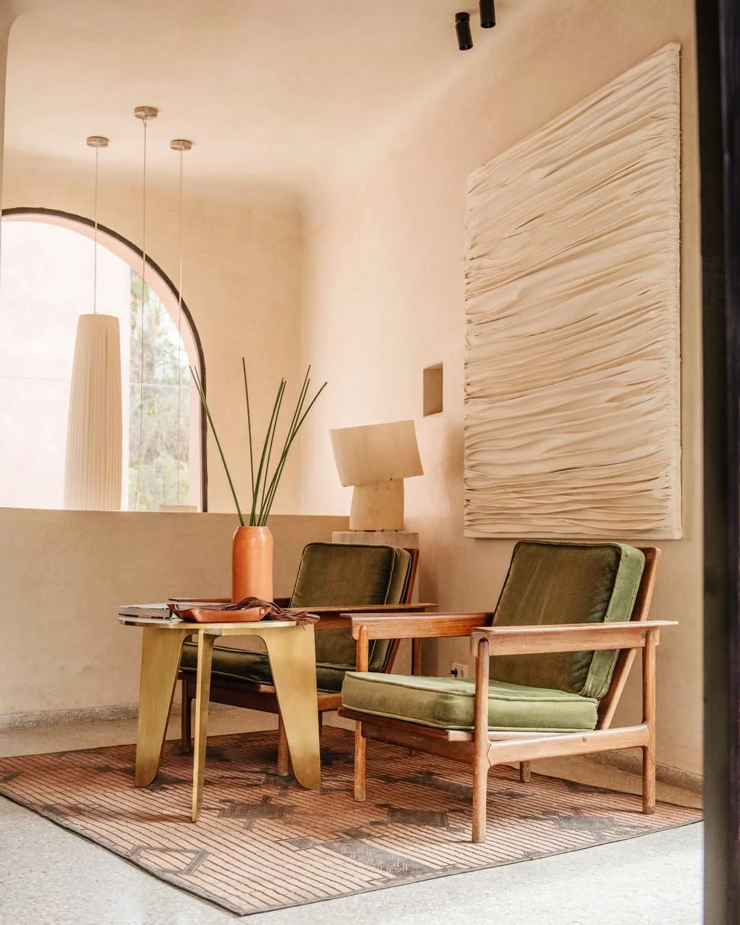 A cozy interior space with two mid-century modern armchairs with green upholstery, a small wooden side table, a tan vase with long reed-like plants, a beige table lamp, and a large textured white wall art piece, with natural light coming through an arched window.